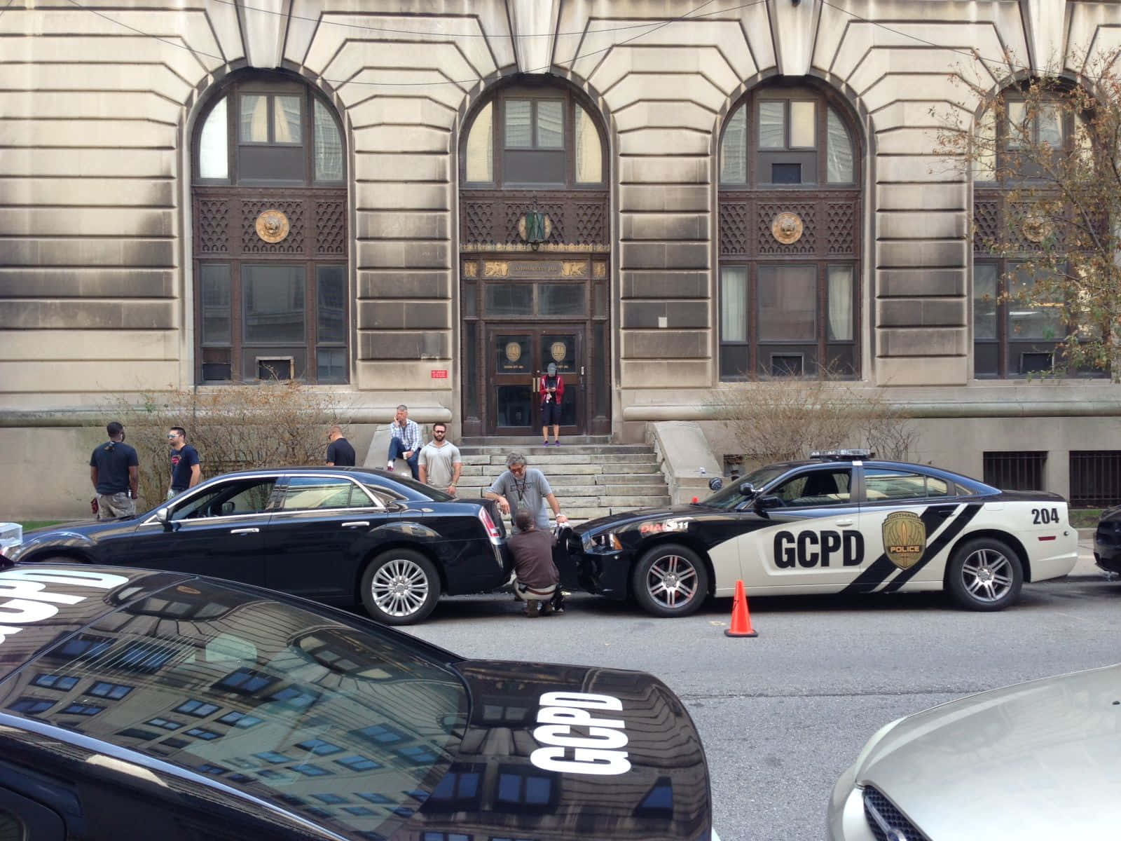 Gotham City Police Department 桌布 壁紙 — 一輛警車和一輛黑色轎車停在一座宏偉的石建築前，人們聚集在車輛周圍。