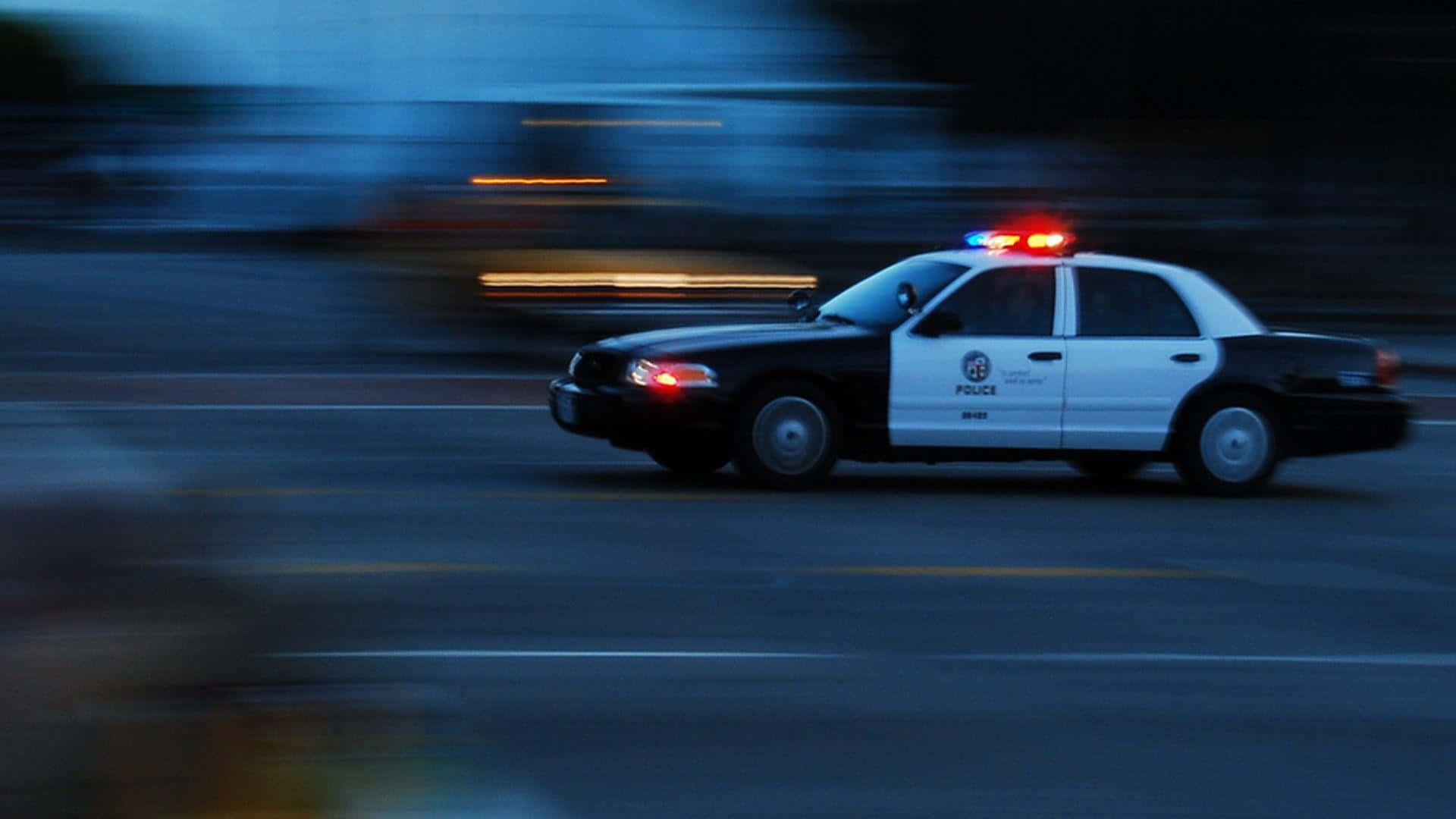 Ford Crown Victoria 桌布 壁紙 — 一輛警車在夜晚穿過模糊的城市街道，閃爍著紅色和藍色的燈光。
