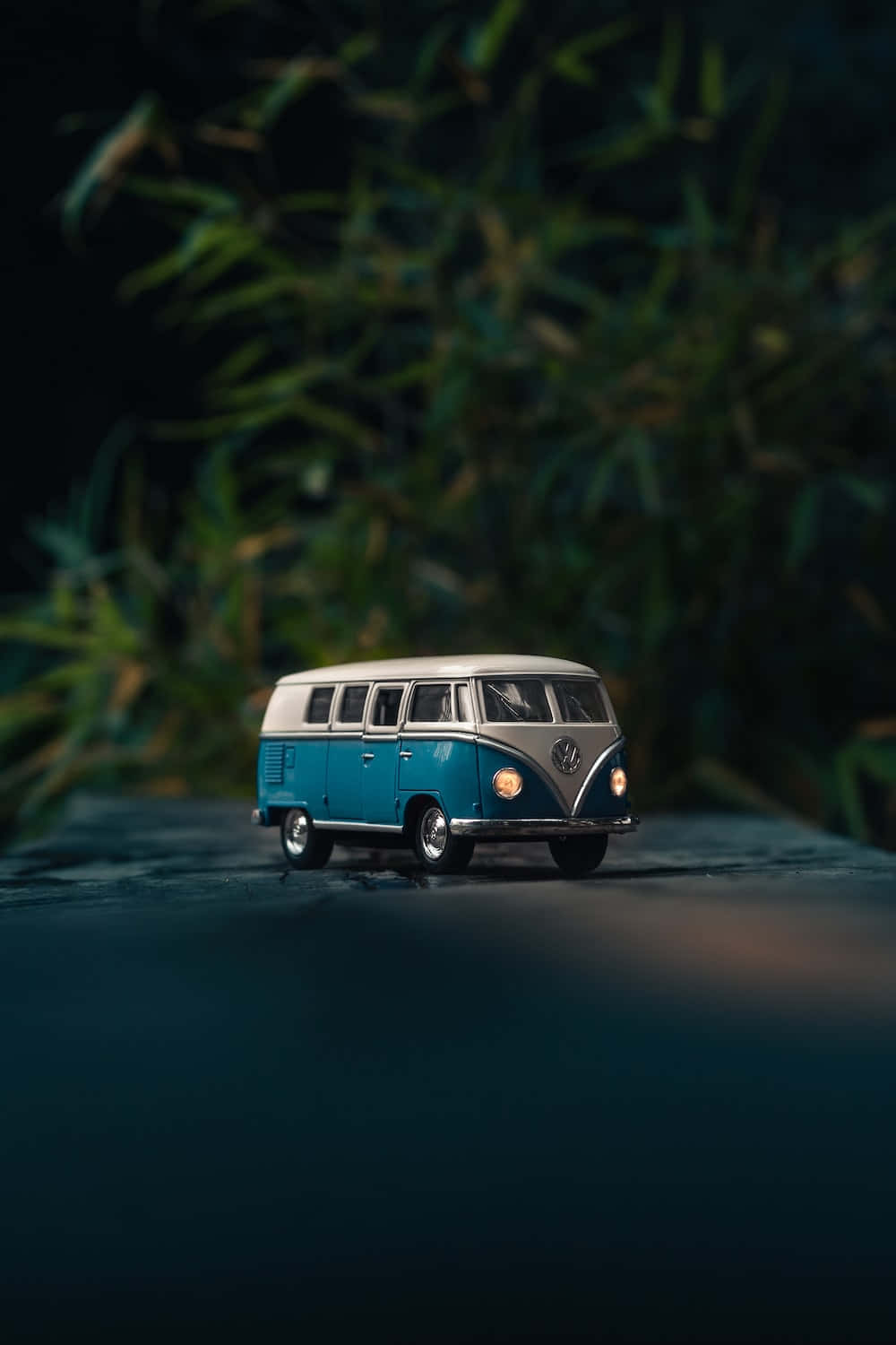 Van 桌布 壁紙 — 一輛迷你藍白復古福斯麵包車停在深色表面上，背後有模糊的綠色樹葉。
