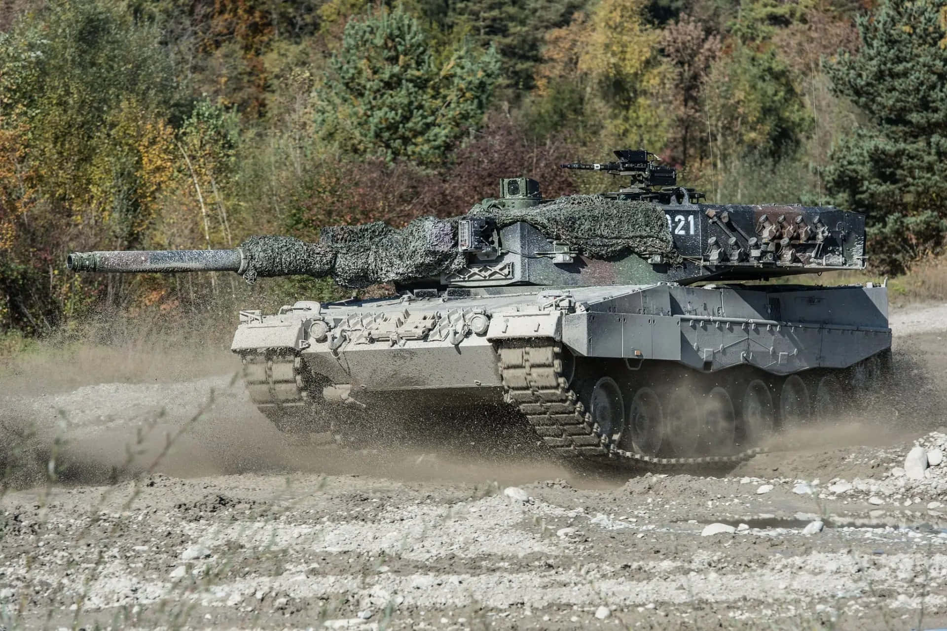 Tanks 桌布 壁紙 — 一輛迷彩軍用坦克在崎嶇的泥土路上行駛，揚起灰塵，穿過秋日森林景觀。