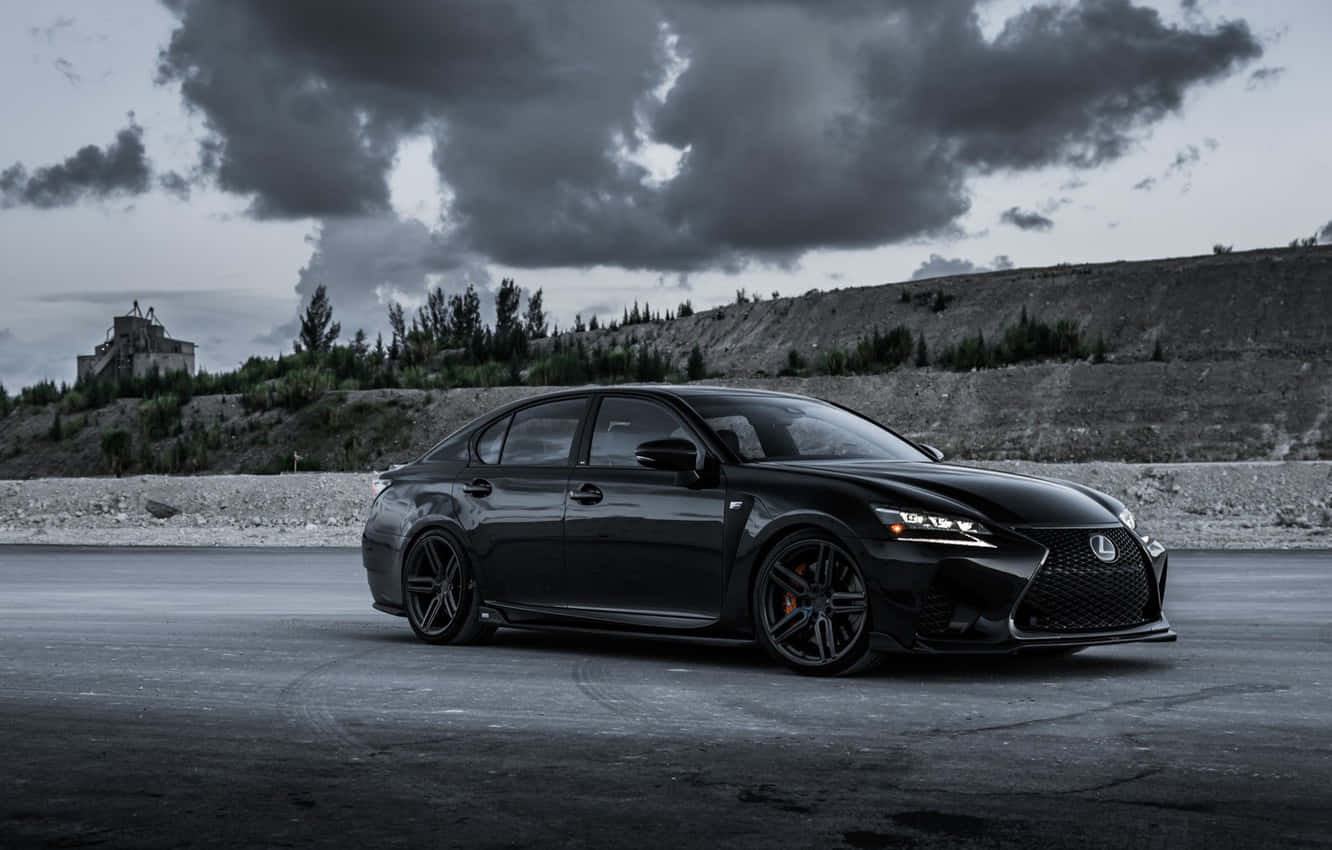Lexus Gs F 桌布 壁紙 — 一輛配備定制輪圈的時尚黑色Lexus轎車，在戲劇性的暴風雲下，停泊在濕漉漉的瀝青路上，遠處山丘上有一座城堡。