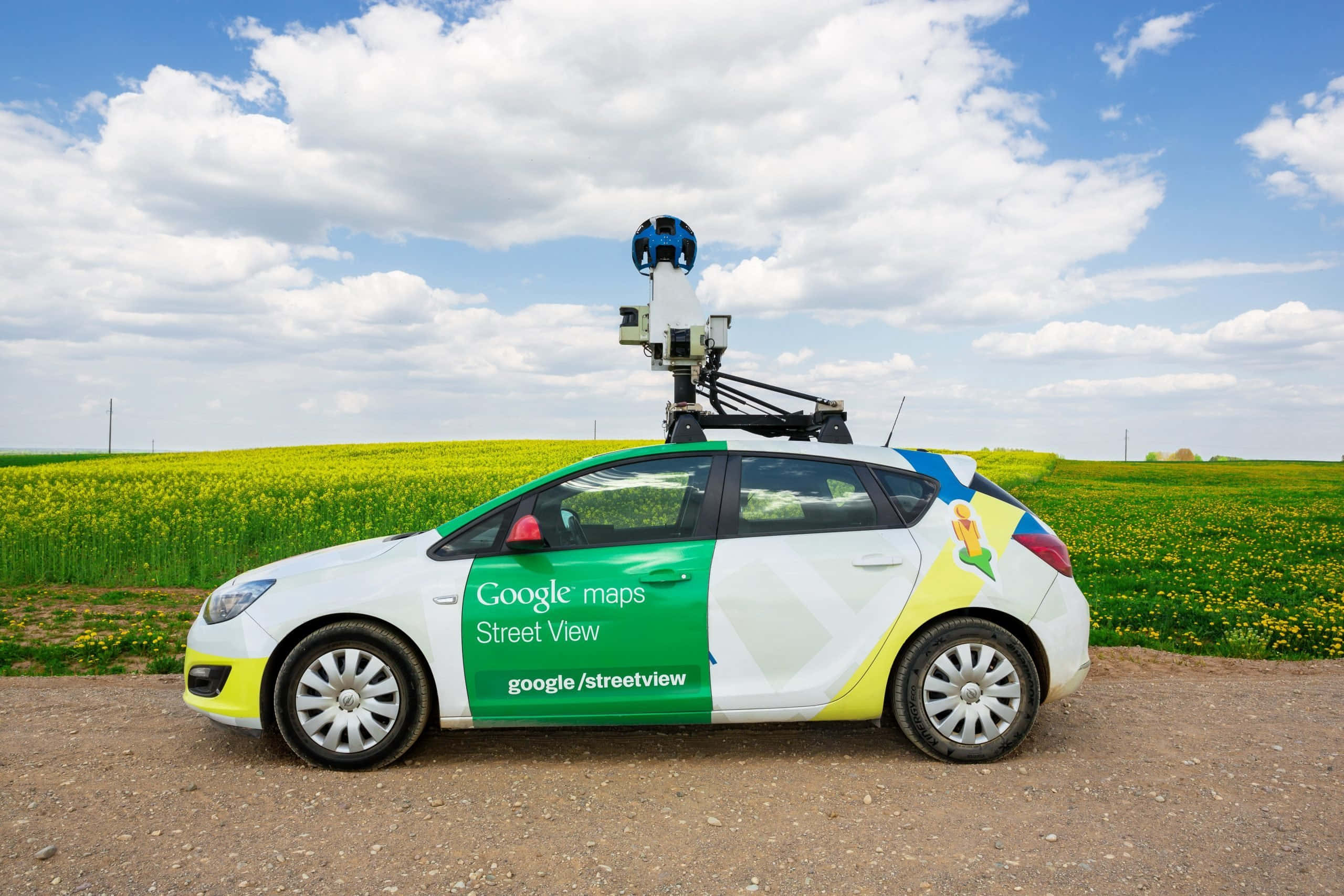 google 桌布 壁紙 — 一輛配備攝影機支架的 Google 街景車停在泥路旁，旁邊是鮮黃色的油菜花田，天空多雲帶藍。
