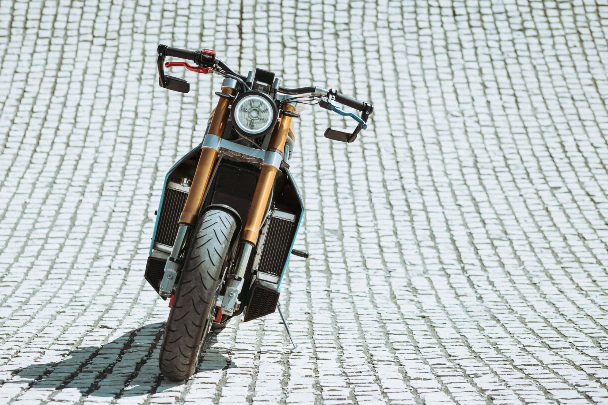Electric Bike 桌布 壁紙 — 一輛配備金色前叉和黑色飾件的訂製機車，停放在陽光普照的鵝卵石街道上，投下清晰的陰影。