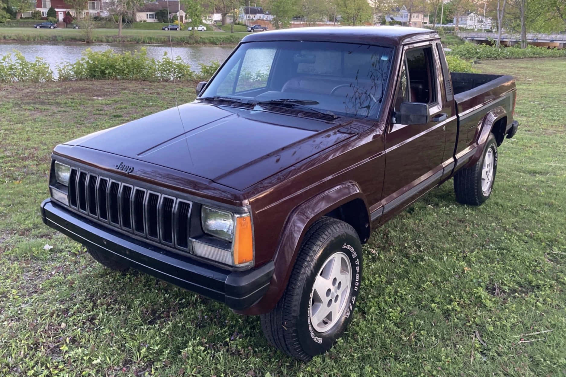 Jeep Comanche 桌布 壁紙 — 一輛酒紅色Jeep Cherokee皮卡車停在平靜湖泊附近的草地上，背景有房屋。