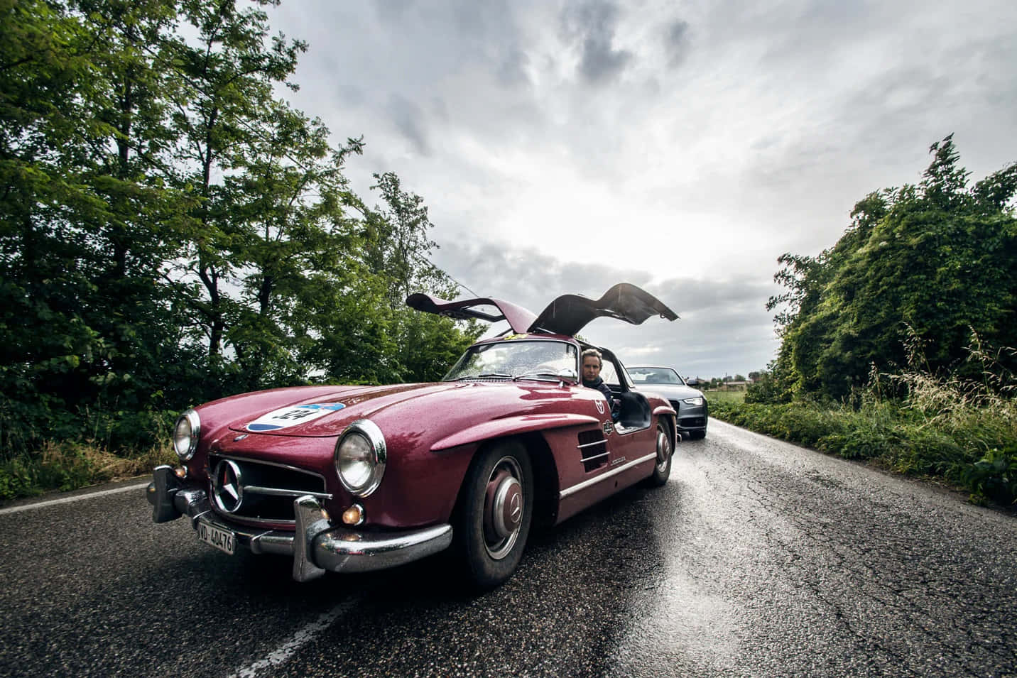 Mercedes 桌布 壁紙 — 一輛酒紅色復古賓士300 SL鷗翼門打開，在潮濕的鄉村道路上行駛，兩旁綠樹成蔭，天空多雲。