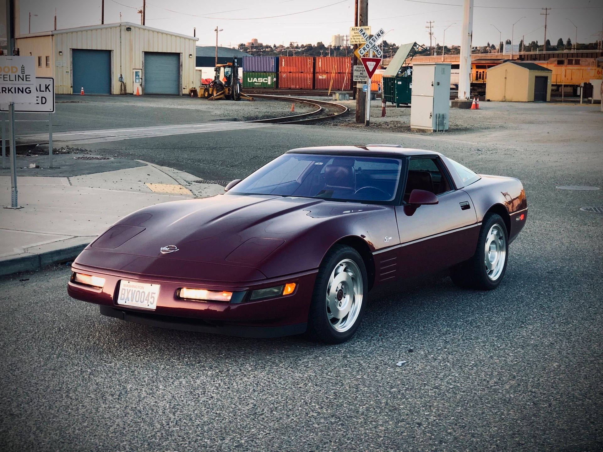 corvette 桌布 壁紙 — 一輛酒紅色的雪佛蘭克爾維特跑車在黃昏時停泊在鐵路軌道和工業建築附近的瀝青路上。