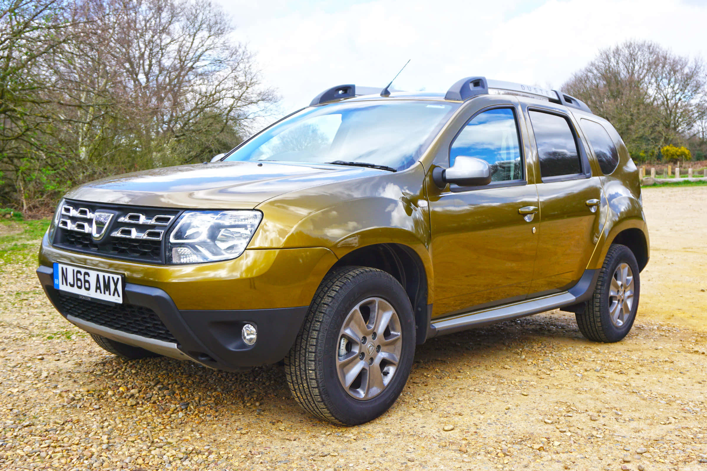Dacia 桌布 壁紙 — 一輛金屬金色的Dacia Duster SUV停放在碎石上，背景是光禿禿的樹木和籬笆，天空多雲。