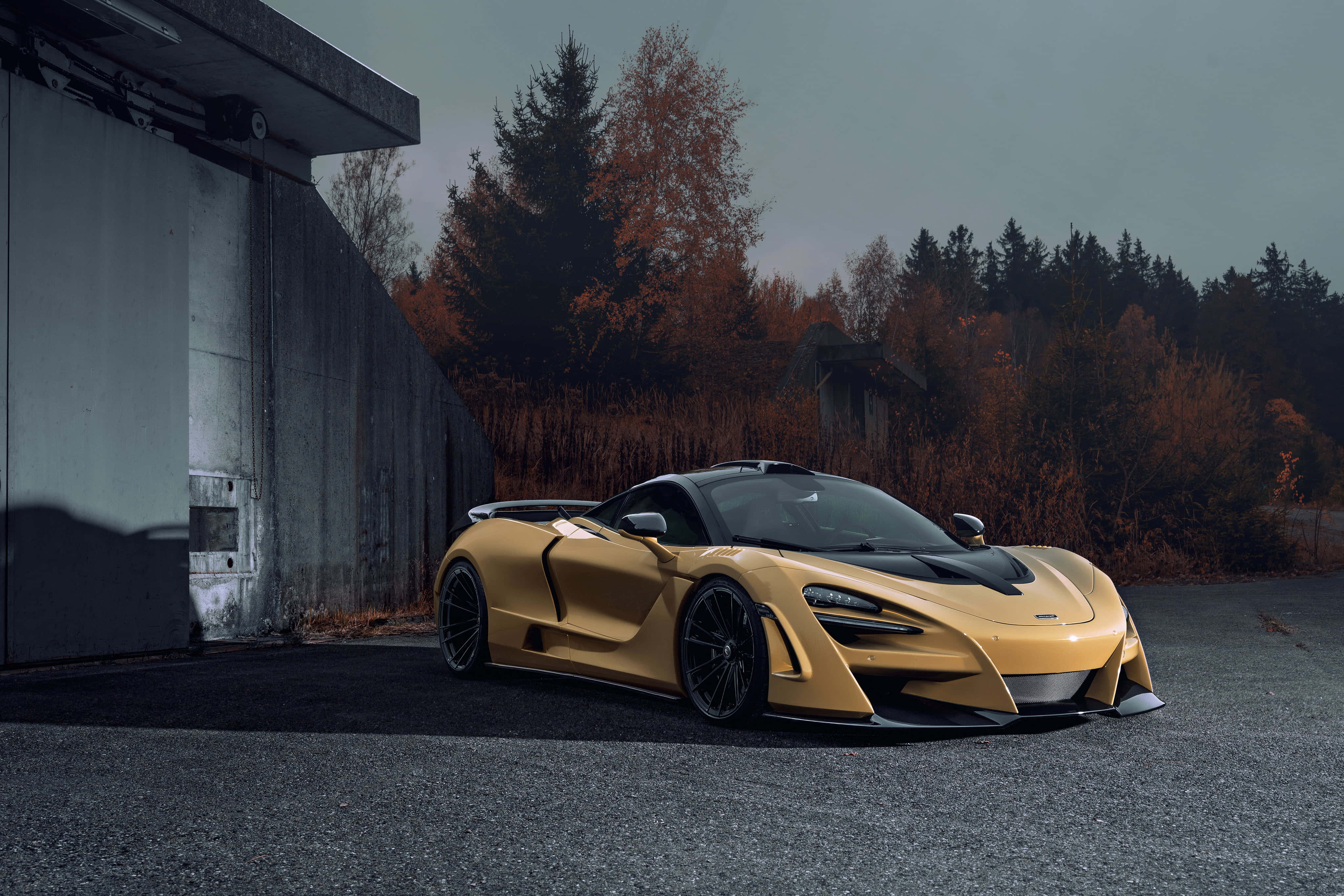 Mclaren 720s 桌布 壁紙 — 一輛金色McLaren跑車停泊在混凝土建築物旁邊，背景有秋天的樹木，天空多雲。