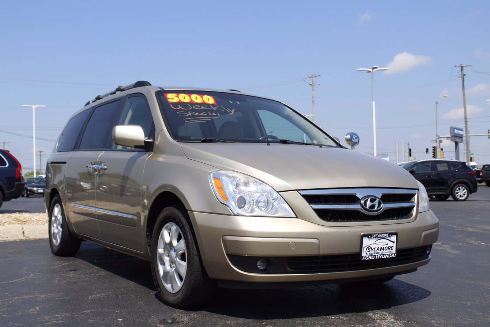 Hyundai Entourage 桌布 壁紙 — 一輛金色現代小型貨車停在經銷商場地，天空清澈湛藍，擋風玻璃上貼有「5000 Weekly Special」標誌。