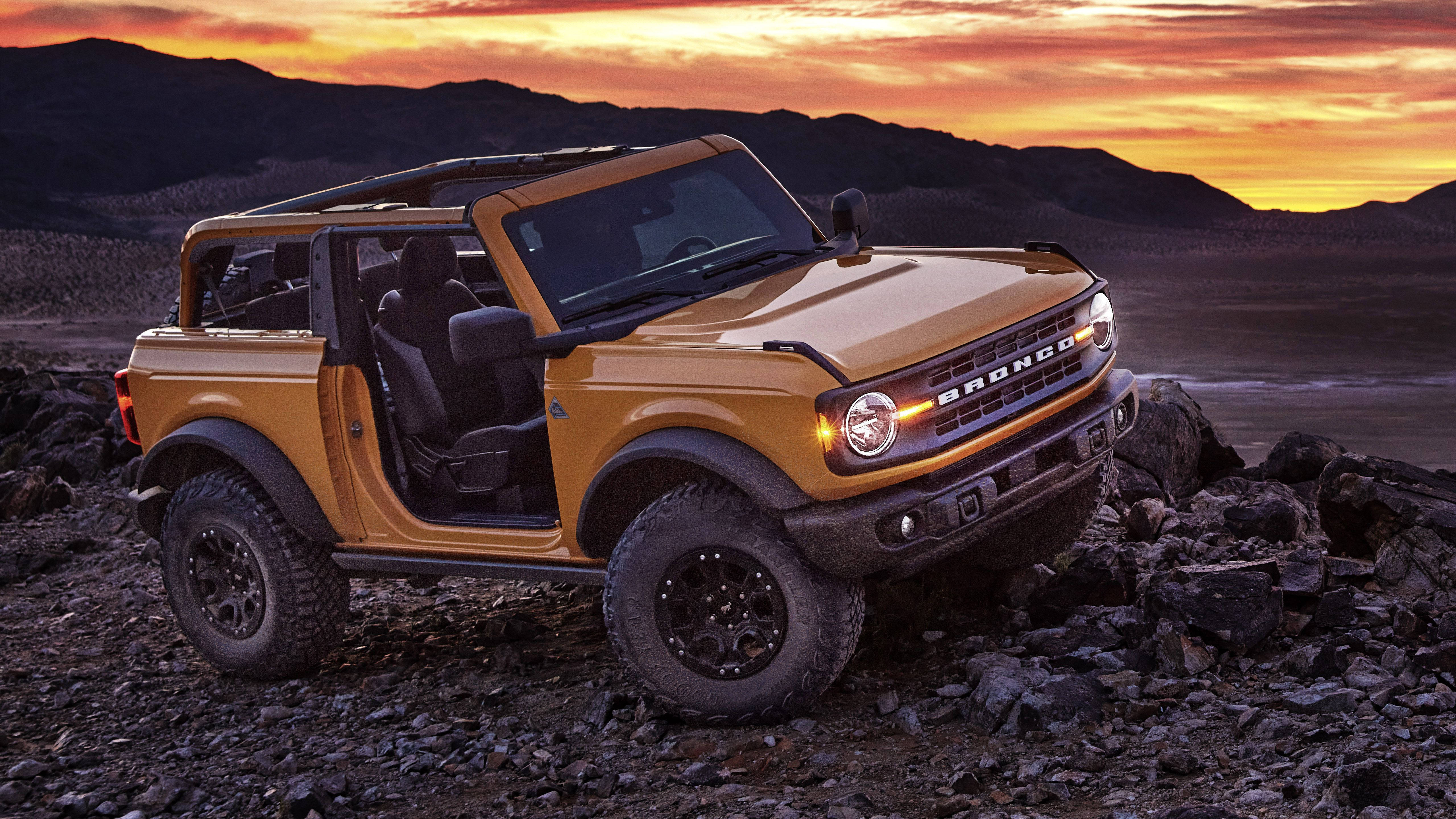 Ford Bronco 桌布 壁紙 — 一輛金色福特Bronco打開車門，停在日落時分的岩石地形上，背景有山脈。