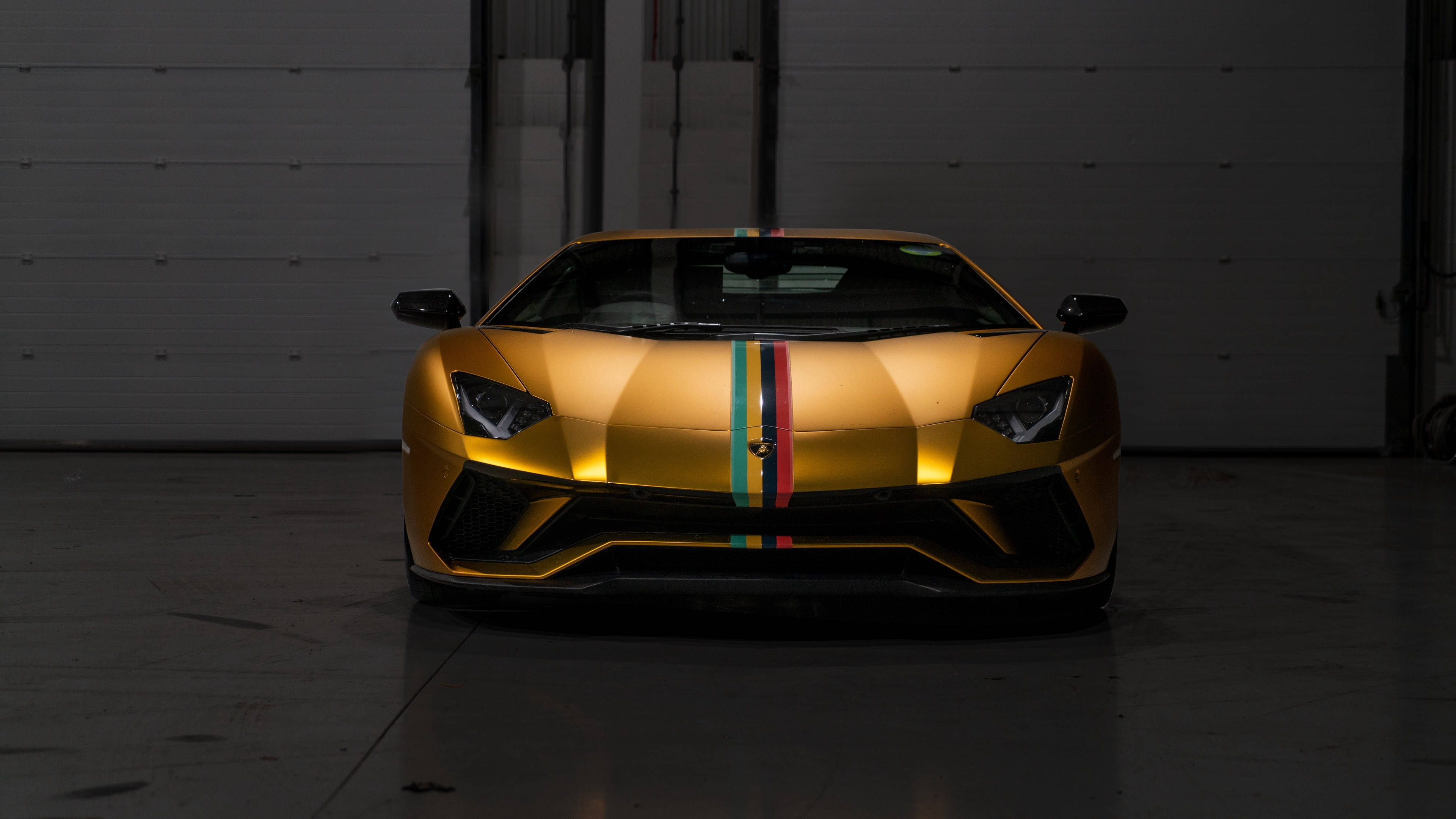 Auto Racing 桌布 壁紙 — 一輛金色藍寶堅尼Aventador帶有彩虹賽車條紋，在昏暗的工業車庫中於聚光燈下閃閃發光。