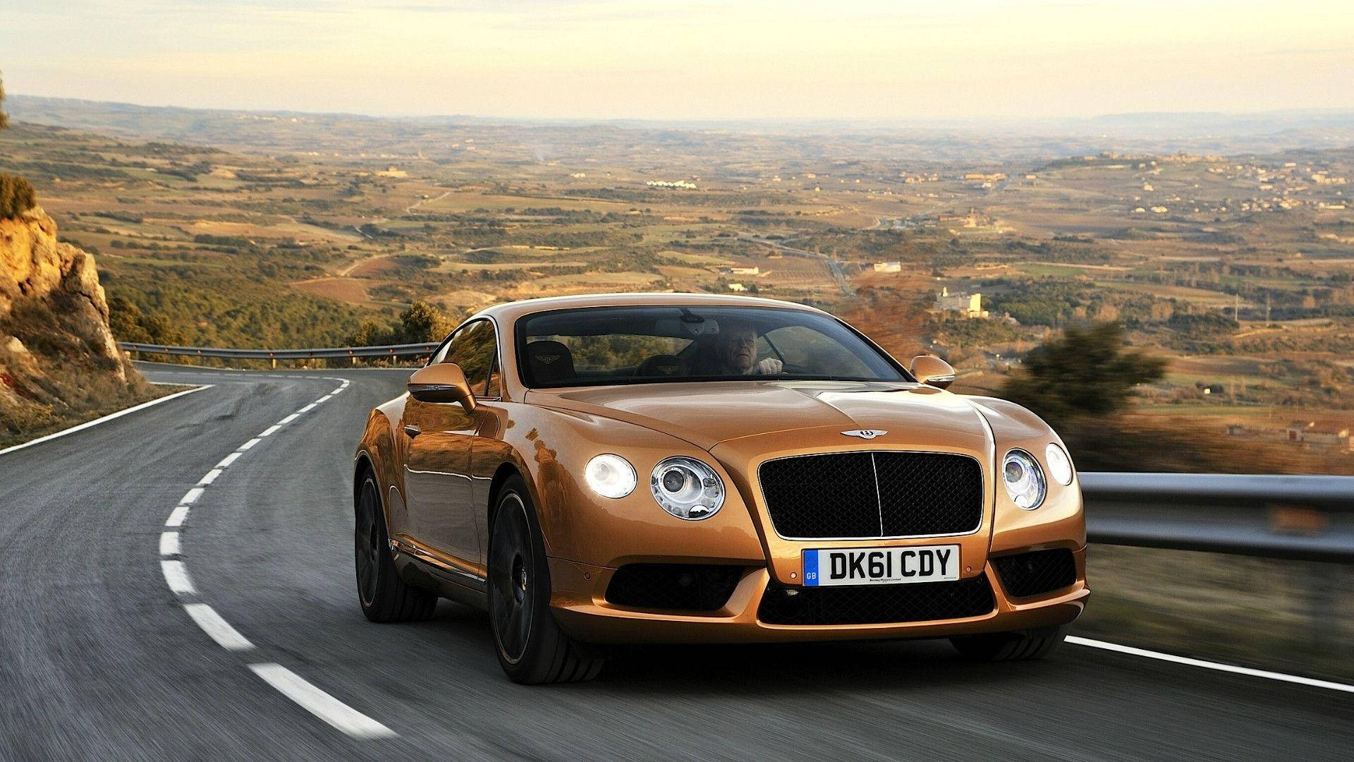 Road 桌布 壁紙 — 一輛金色賓利 Continental GT 在蜿蜒的山路上疾馳，廣闊的丘陵景觀在柔和的金色天空下。