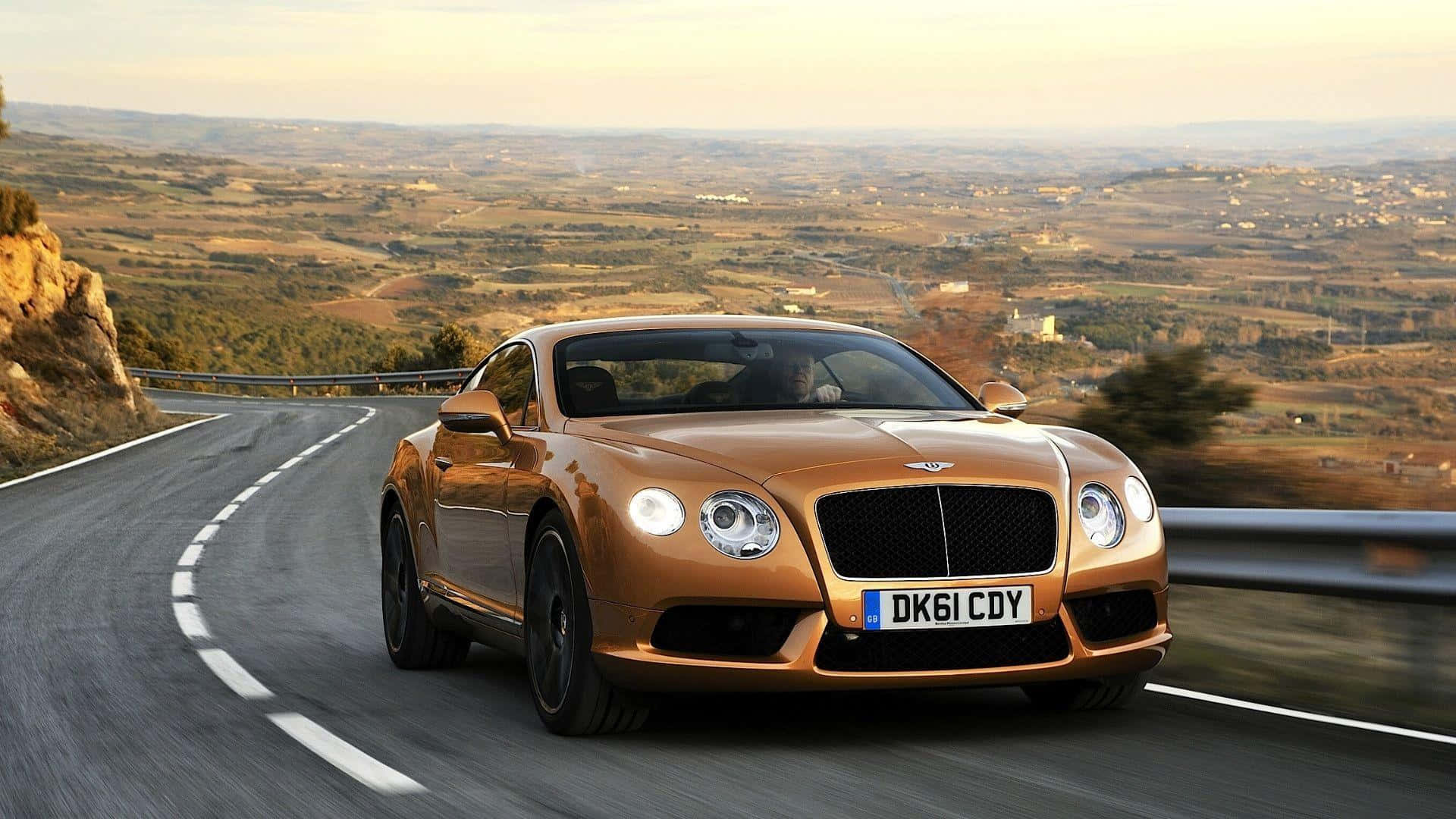 Best Bentley 桌布 壁紙 — 一輛金色賓利 Continental GT 沿著蜿蜒的山路疾馳，廣闊的丘陵景觀在柔和的金色天空下展開。
