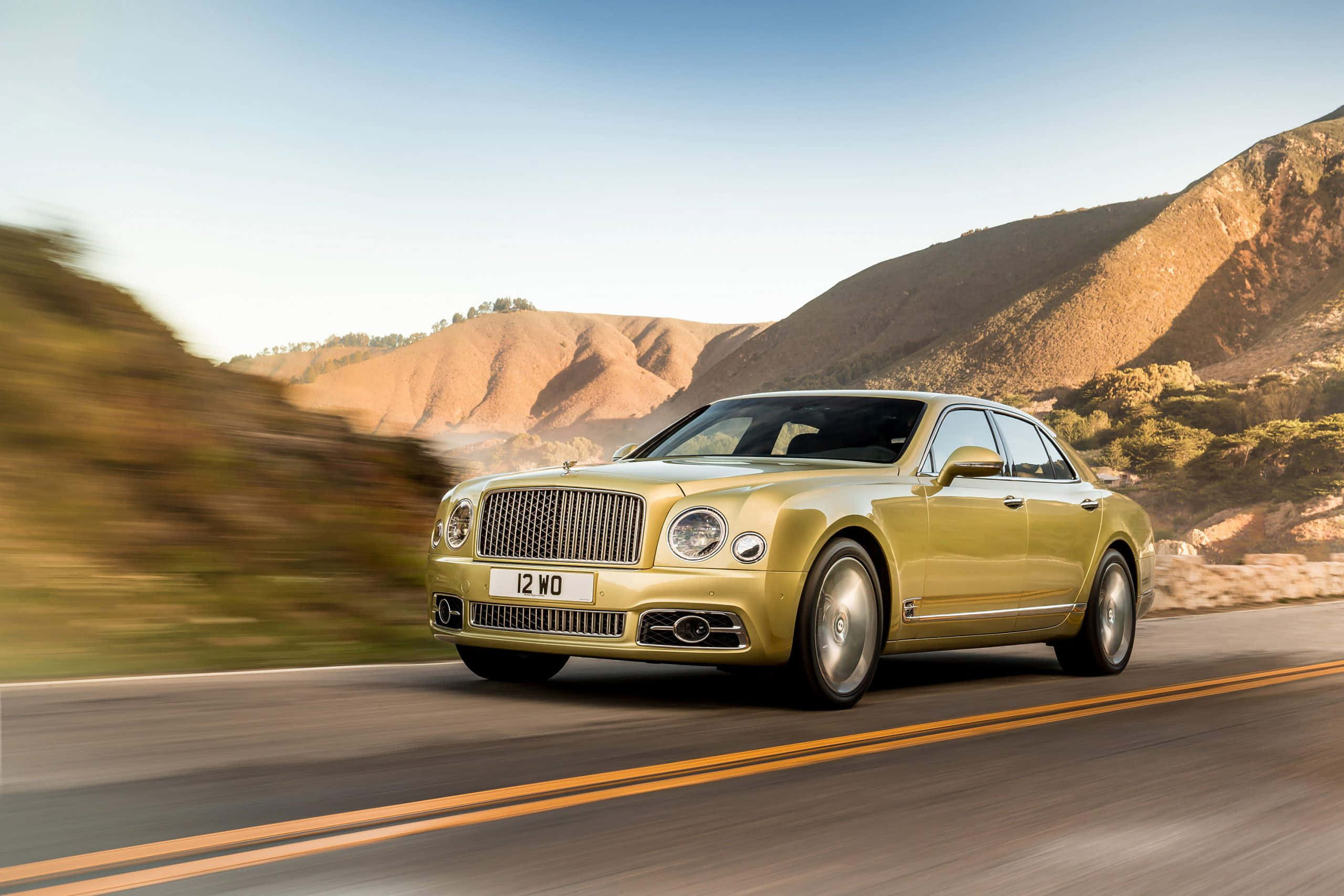 Bentley Mulsanne 桌布 壁紙 — 一輛金色賓利Mulsanne在晴朗藍天下沿蜿蜒山路疾馳，風景模糊。