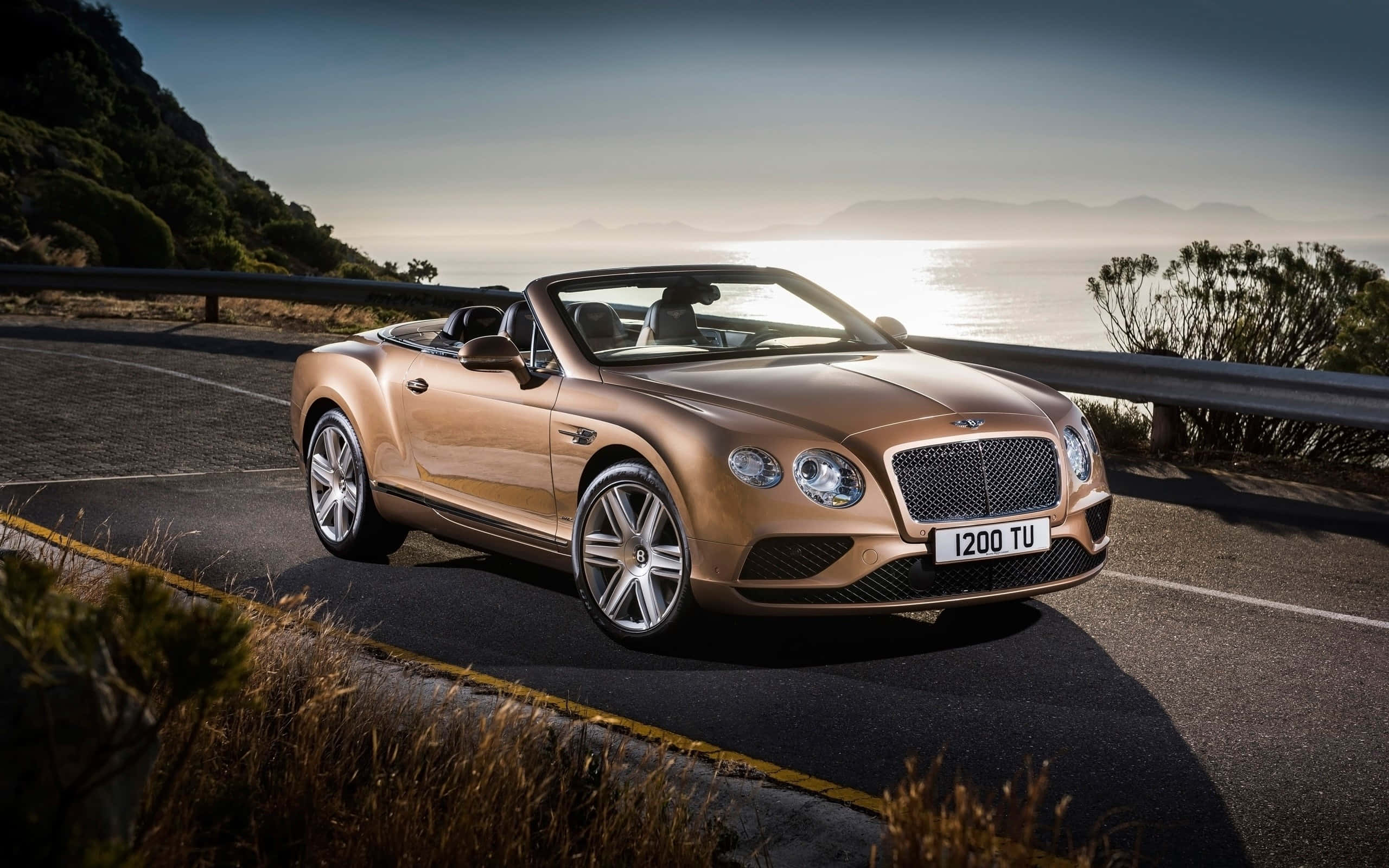 Bentley Continental Gt Convertible 桌布 壁紙 — 一輛金色賓利敞篷車沿著海岸公路行駛，背景是山脈和海洋，正值日出時分。