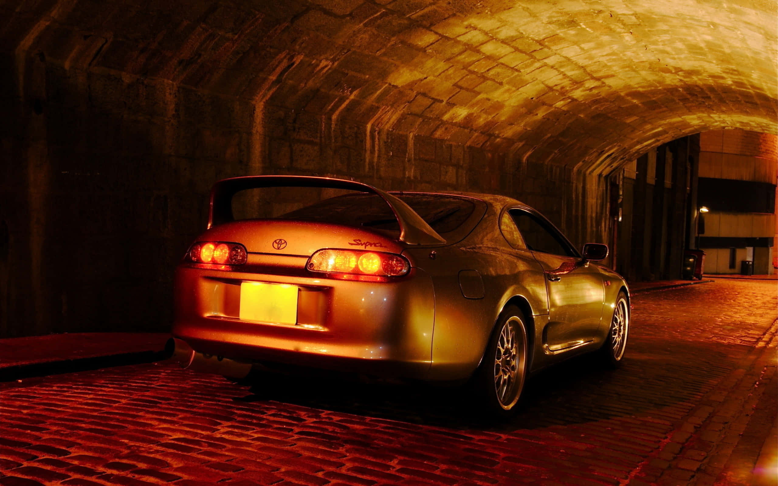 Supra 桌布 壁紙 — 一輛金色跑車帶有發光的尾燈，停放在光線昏暗的石頭隧道內，地面鋪有紅磚。