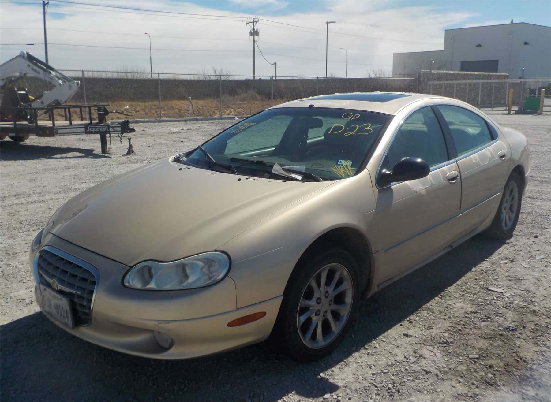 Chrysler Lhs 桌布 壁紙 — 一輛金色轎車停泊在圍欄工業區的碎石地上，上方是明亮的白天天空。