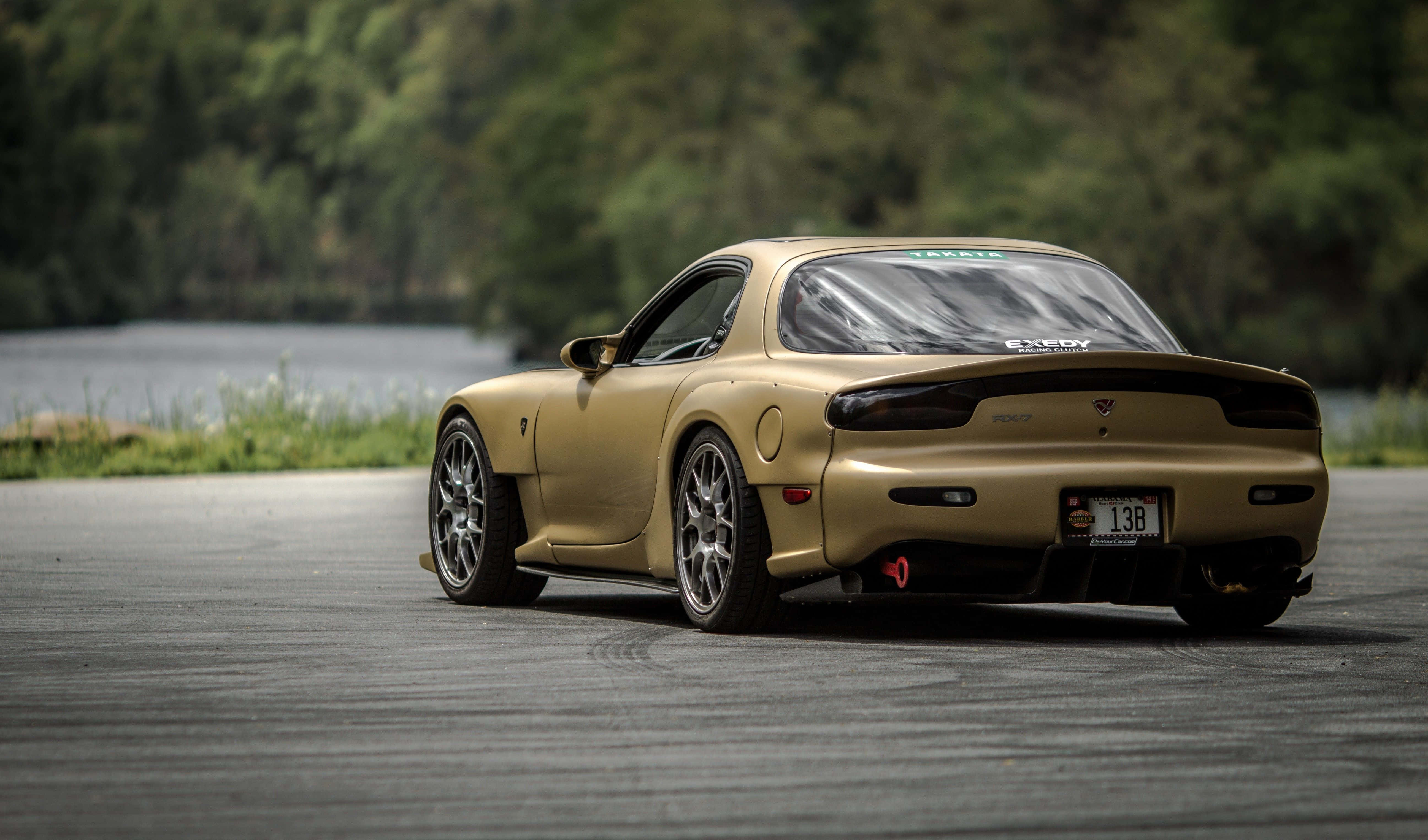 Mazda 桌布 壁紙 — 一輛金色馬自達RX-7跑車停放在鋪砌的道路上，旁邊是一個平靜的湖泊，背景是樹木繁茂的山丘。