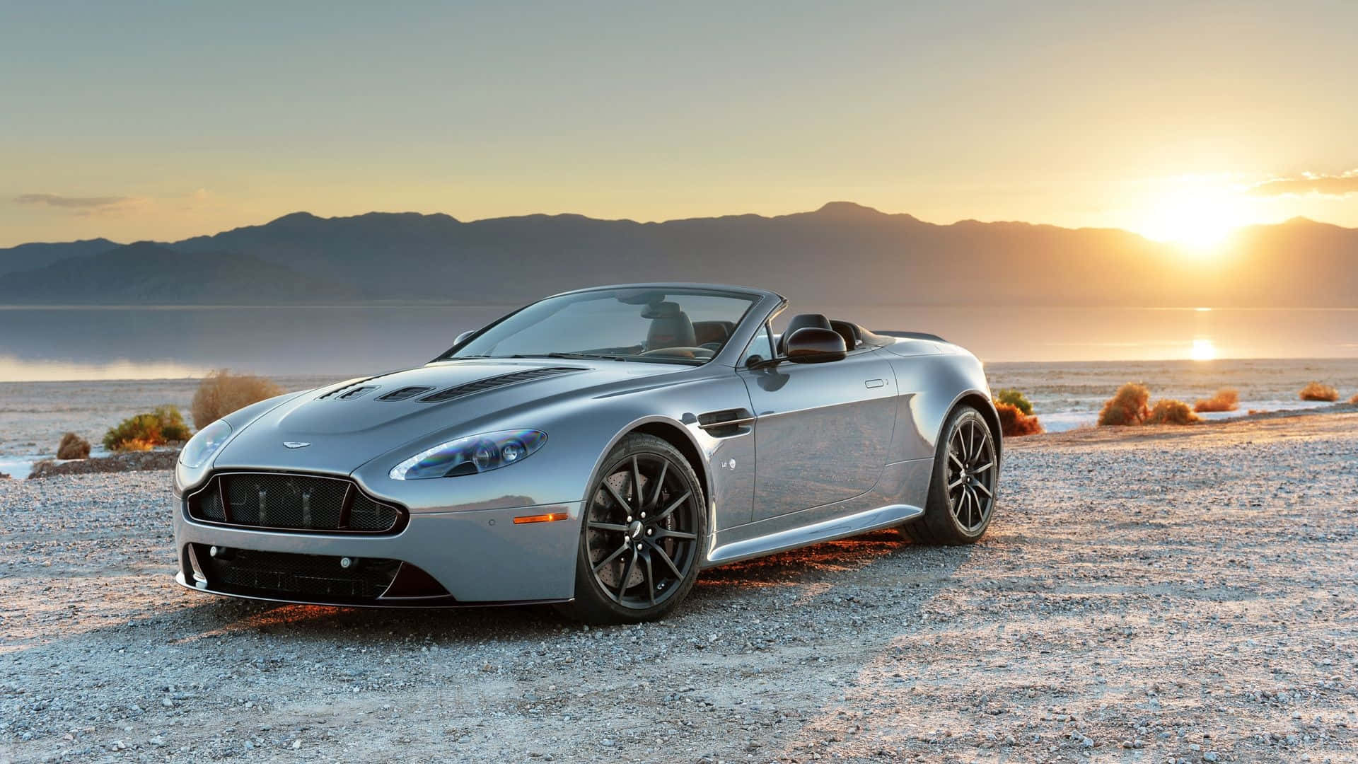 Aston Martin V12 Vantage 桌布 壁紙 — 一輛銀色 Aston Martin V12 Vantage 敞篷車停在日落時分的碎石沙漠海岸上，背景有山脈。