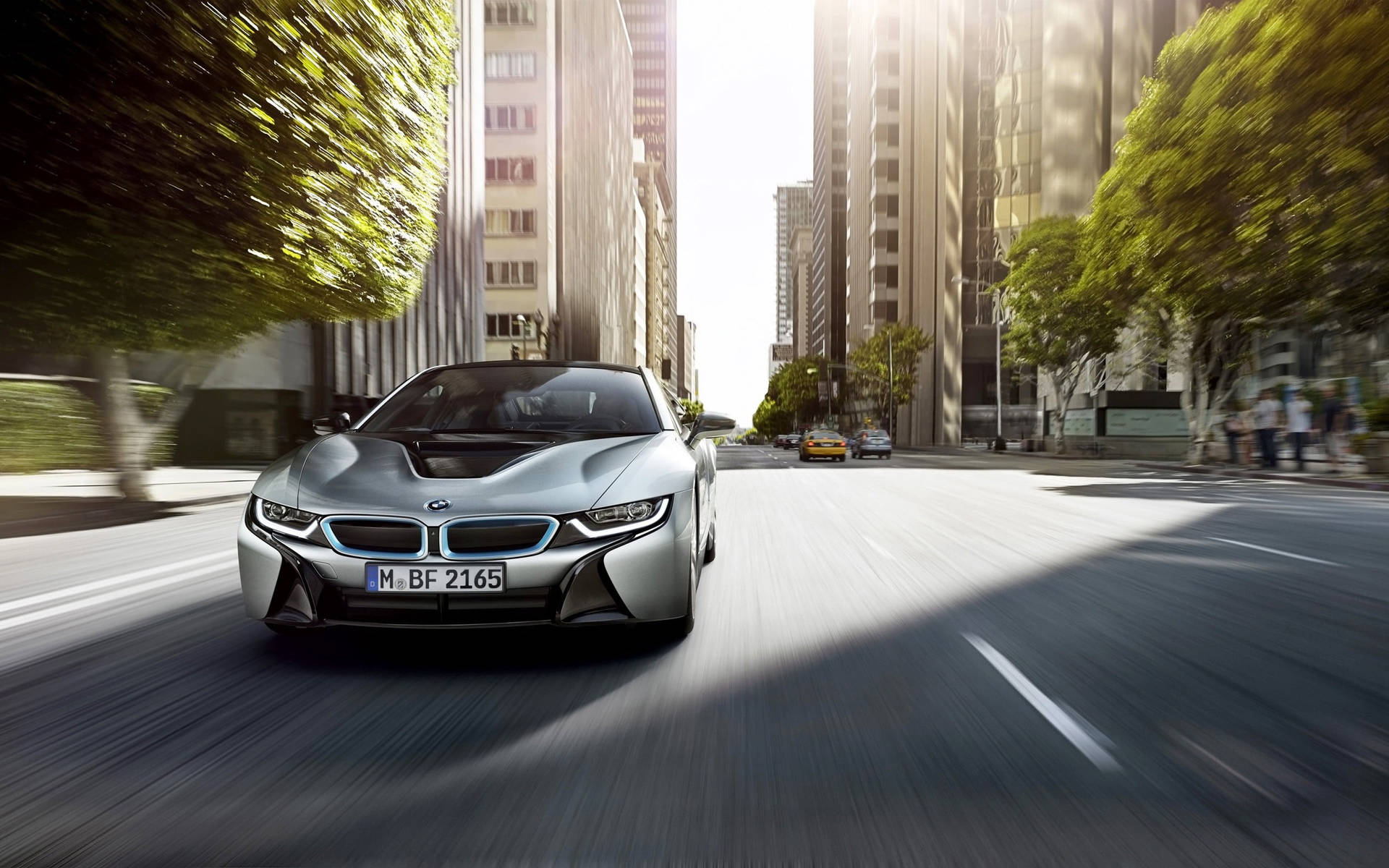BMW 動圖 桌布 壁紙 — 一輛銀色 BMW i8 跑車在陽光明媚、兩旁高樓大廈和綠樹成蔭的城市街道上奔馳。