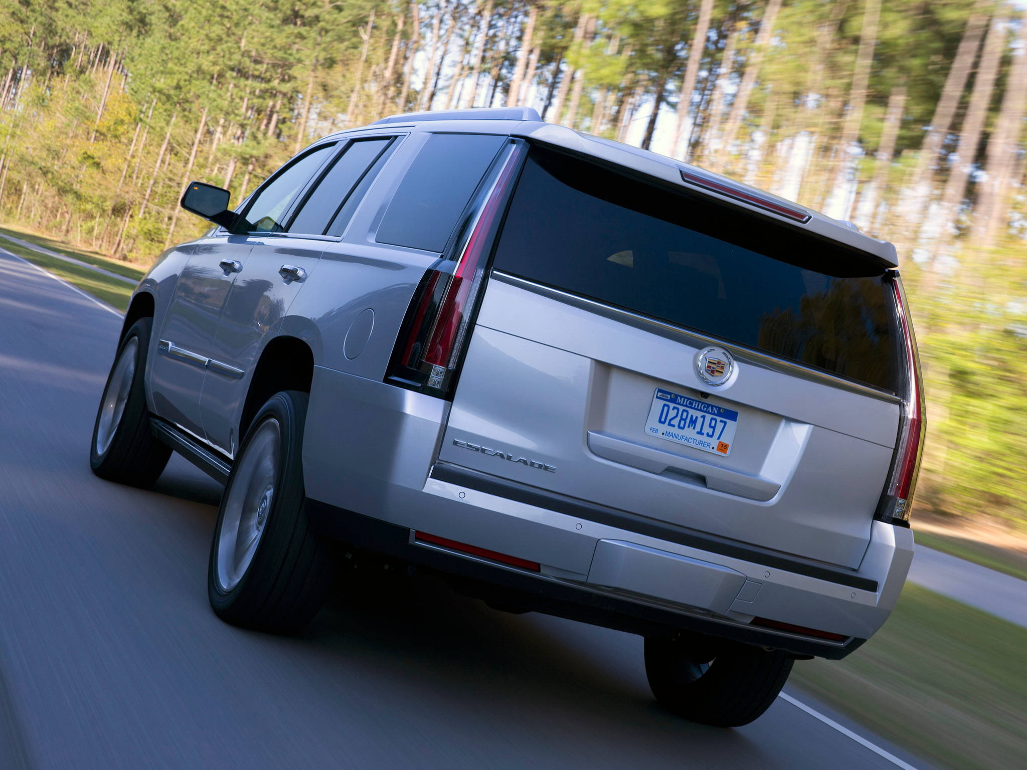 凱迪拉克 iPhone 桌布 壁紙 — 一輛銀色 Cadillac Escalade SUV 在陽光明媚的森林中沿著蜿蜒的道路疾馳，背景中的樹木模糊不清。