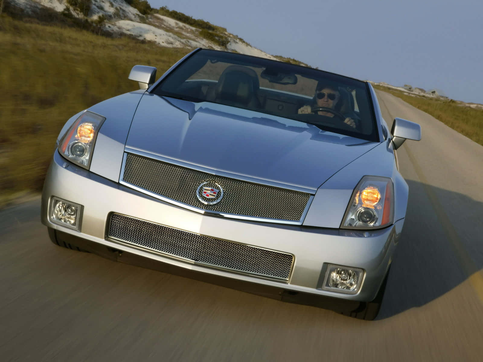Cadillac Xlr 桌布 壁紙 — 一輛銀色 Cadillac 敞篷車沿著蜿蜒的海岸道路疾駛，背景是模糊的草地山丘，天空清澈湛藍。