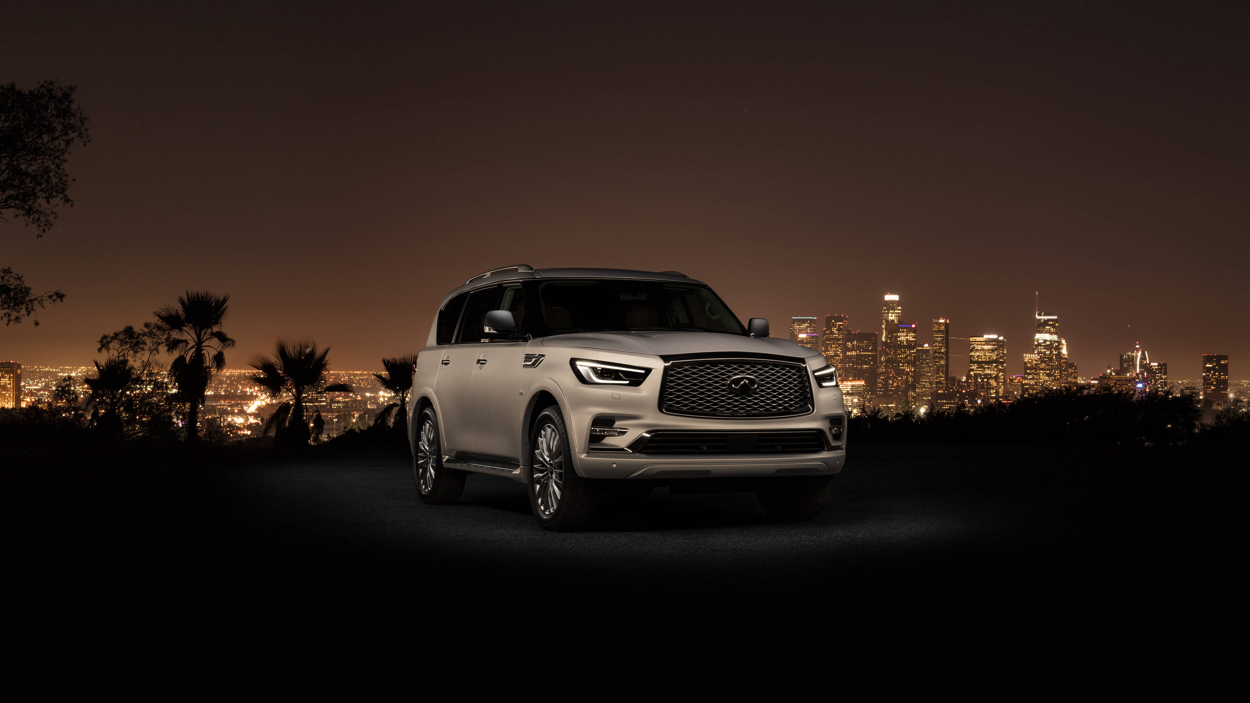 Infiniti 桌布 壁紙 — 一輛銀色 Infiniti QX80 SUV 停泊在山丘上，俯瞰著夜晚充滿活力的城市天際線，前景有棕櫚樹的剪影。