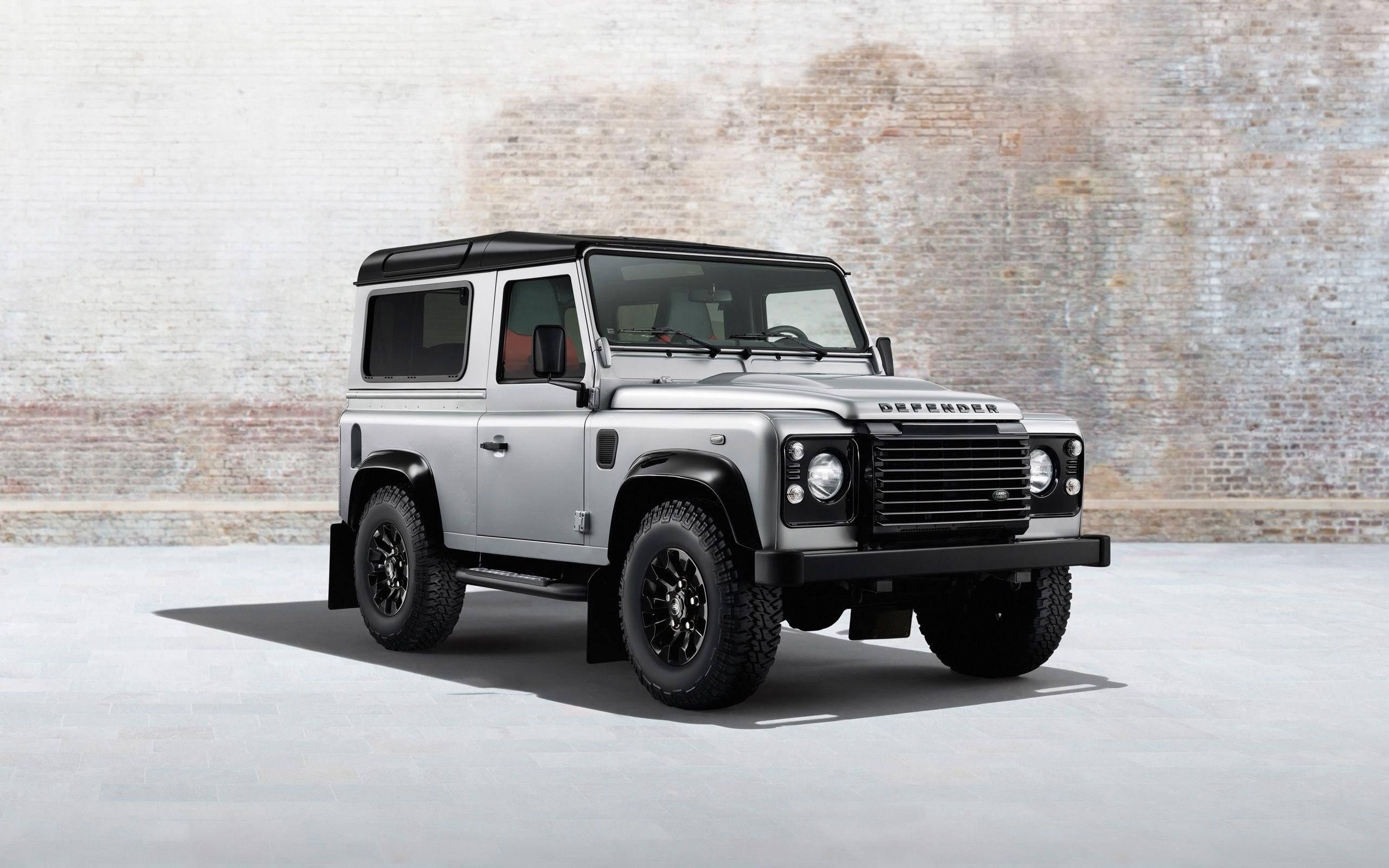 荒原路華 桌布 壁紙 — 一輛銀色 Land Rover Defender 在極簡的工作室環境中，大膽地矗立在風化的磚牆前。