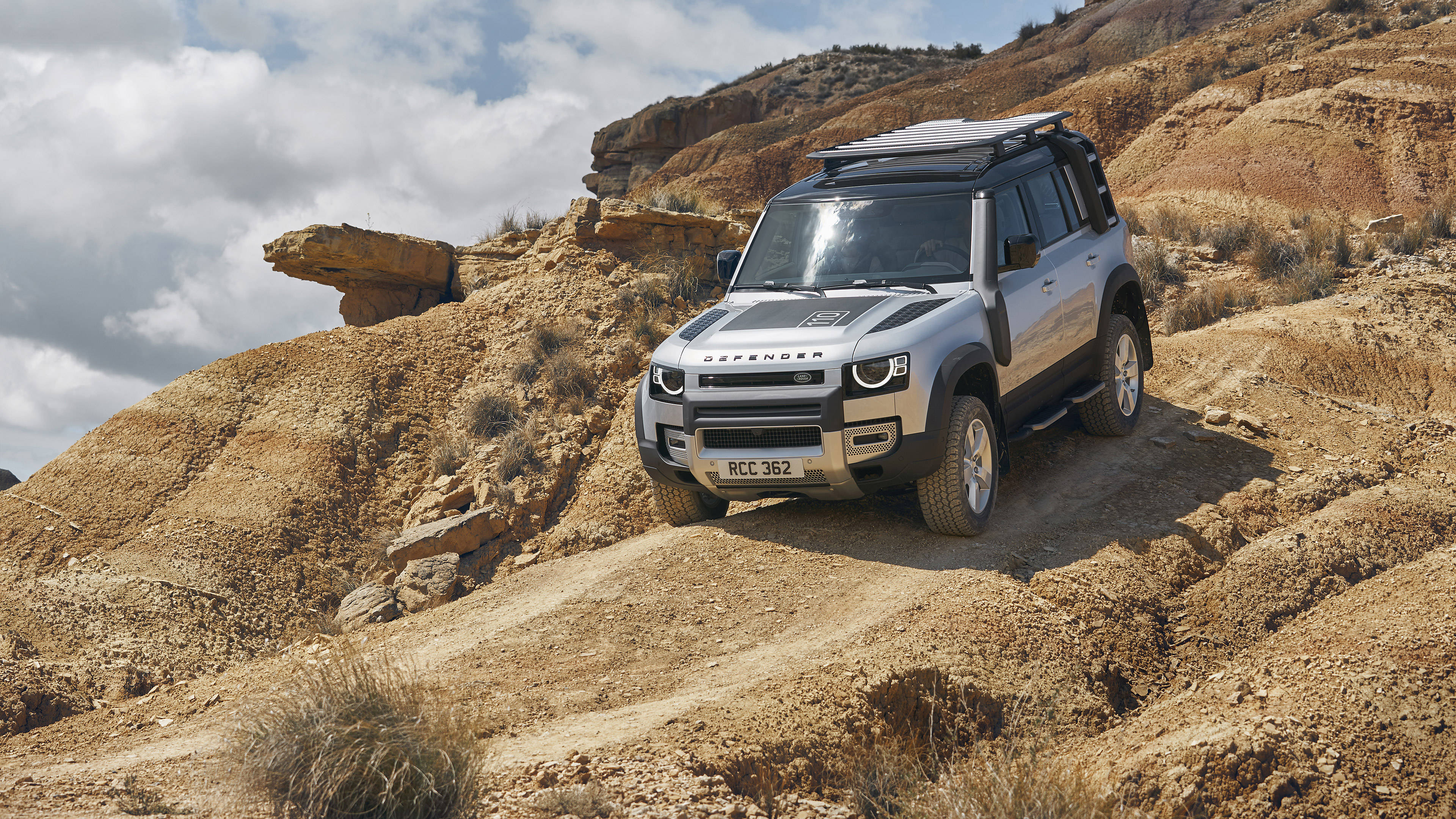 land rover iPhone 桌布 壁紙 — 一輛銀色 Land Rover Defender 在部分多雲的天空下，穿越乾旱岩石地形的崎嶇泥路。