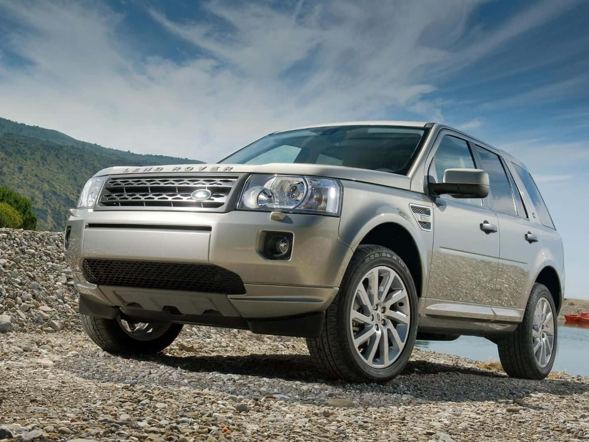 Land Rover Freelander 桌布 壁紙 — 一輛銀色 Land Rover Freelander 停泊在陽光明媚的碎石岸邊，旁邊是平靜的水面，遠處有綠色山丘，天空蔚藍。