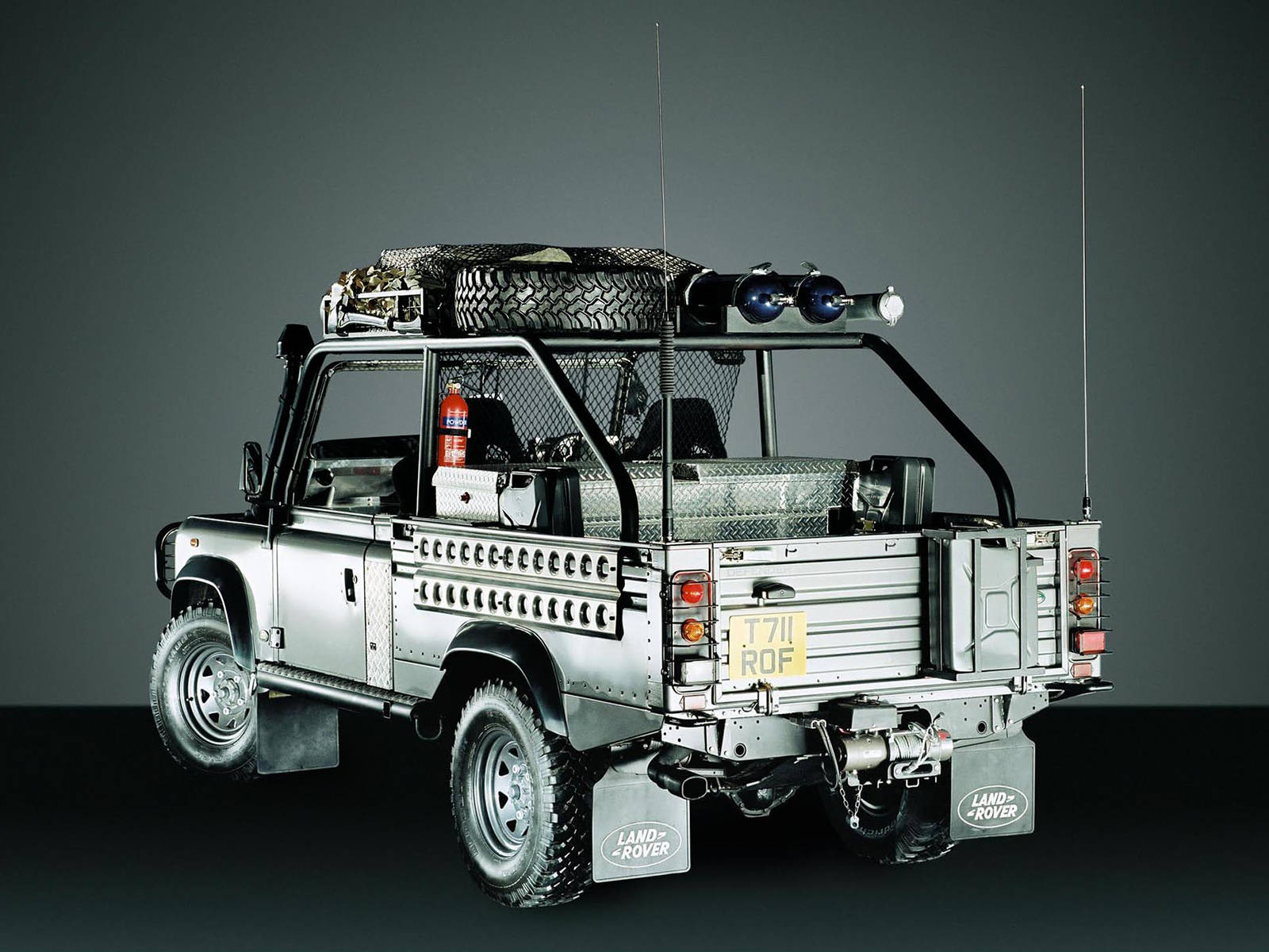 land rover iPhone 桌布 壁紙 — 一輛銀色 Land Rover Defender 皮卡車，配有車頂行李架、備用輪胎和緊急燈，停在深灰色攝影棚背景前。