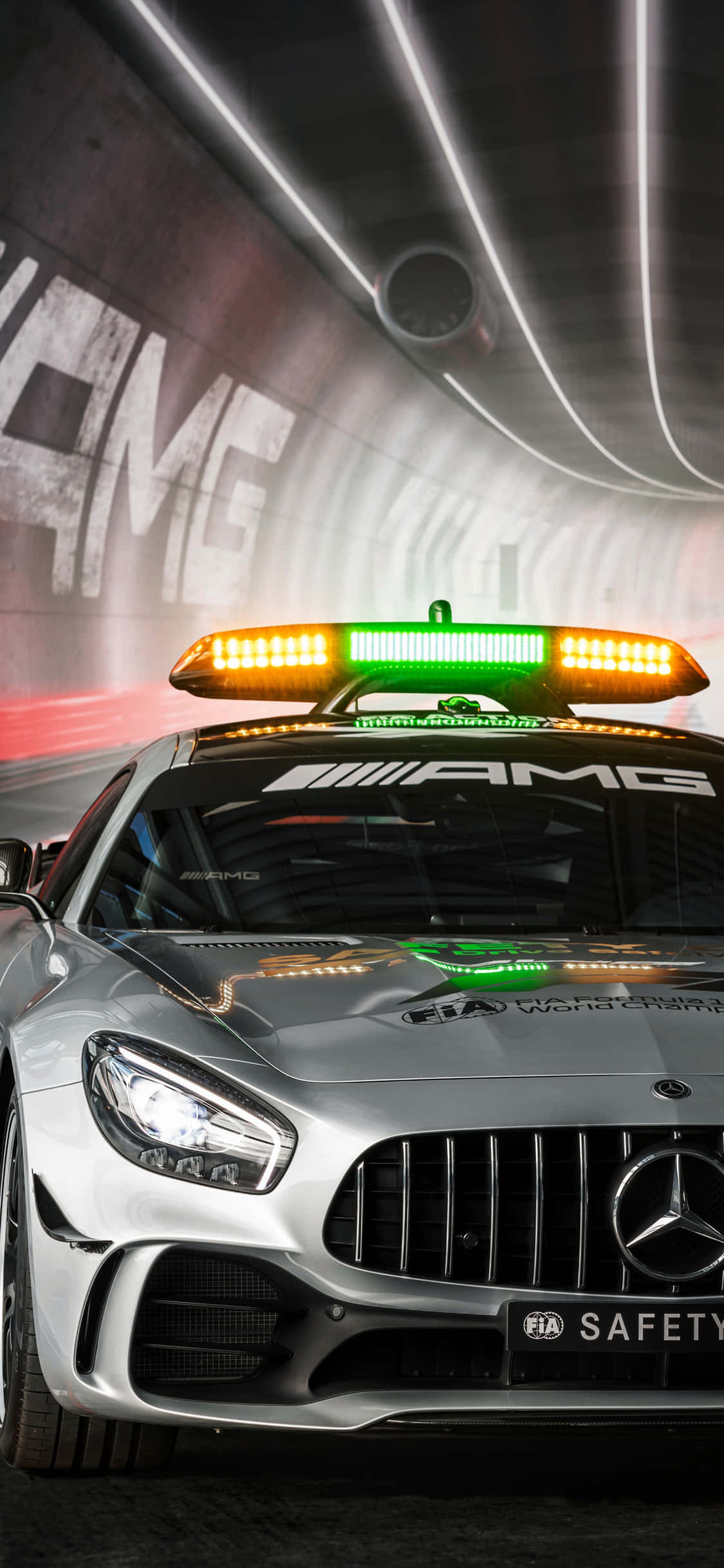 iPhone X Amg Gt-r 桌布 壁紙 — 一輛銀色 Mercedes-AMG 安全車，帶有閃爍的橙色和綠色燈光，在牆上有 'AMG' 塗鴉的隧道中疾馳。