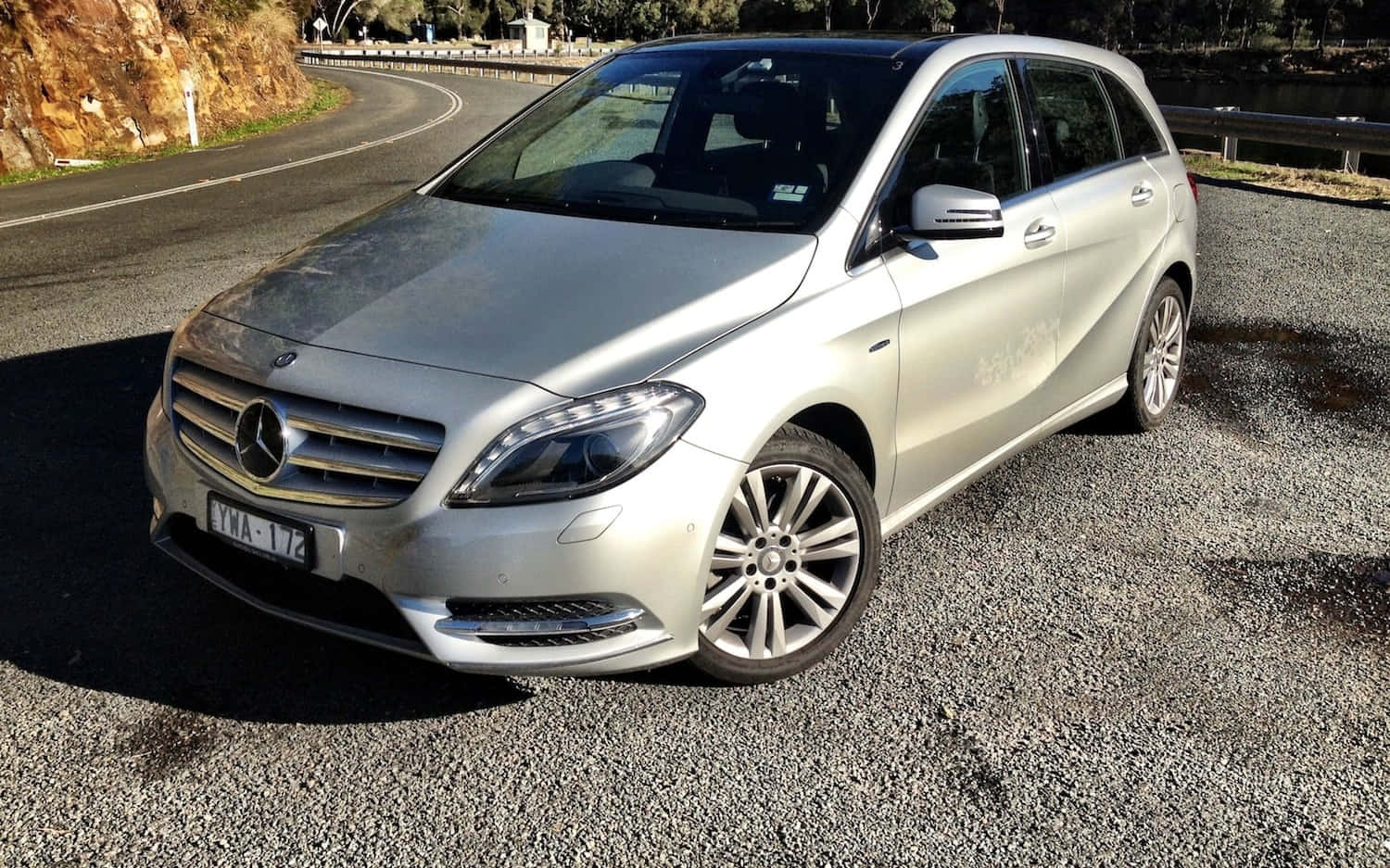 Mercedes Benz B-class 桌布 壁紙 — 一輛銀色 Mercedes-Benz 旅行車停放在碎石路上，旁邊是蜿蜒的道路，背景有樹木和岩石峭壁。