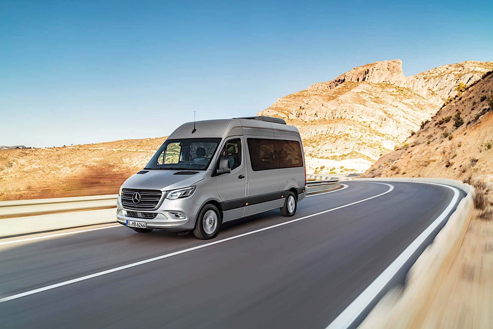 Mercedes Benz Sprinter 桌布 壁紙 — 一輛銀色 Mercedes-Benz Sprinter 客貨車在晴朗藍天下沿著蜿蜒的沙漠公路行駛。