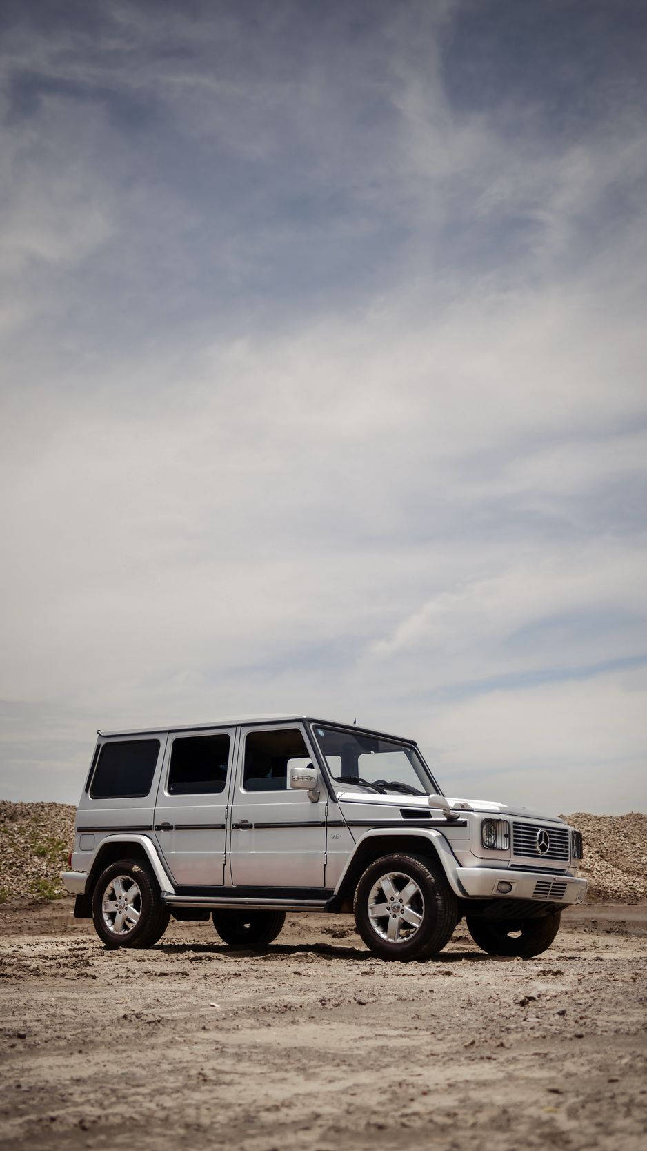 X 桌布 壁紙 — 一輛銀色 Mercedes-Benz G-Class SUV 獨自停在一片荒蕪的泥地上，上方是廣闊的多雲天空。