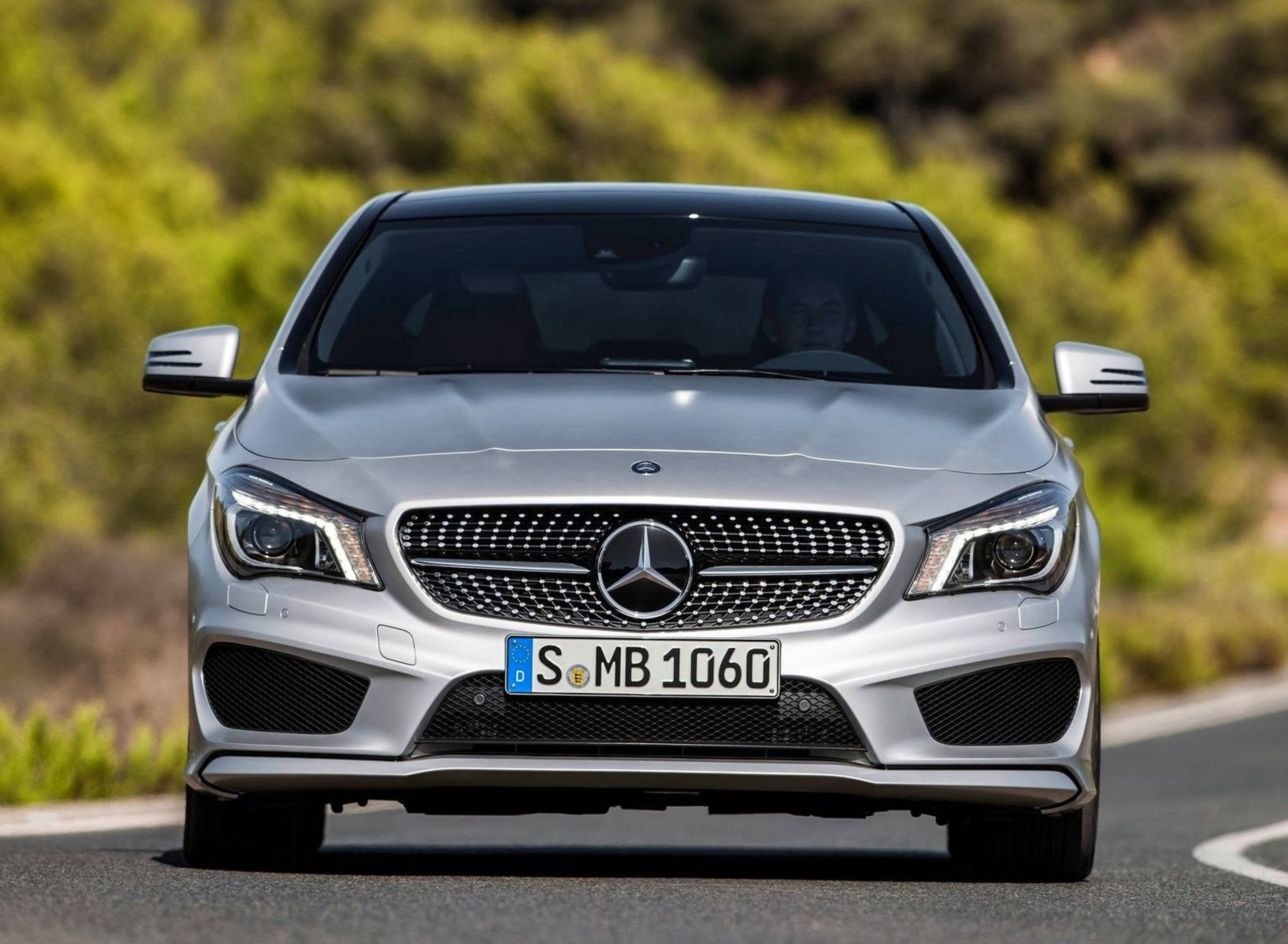 高清 桌布 壁紙 — 一輛銀色 Mercedes-Benz CLA 跑車在蜿蜒的道路上行駛，背景是模糊的綠色樹葉。