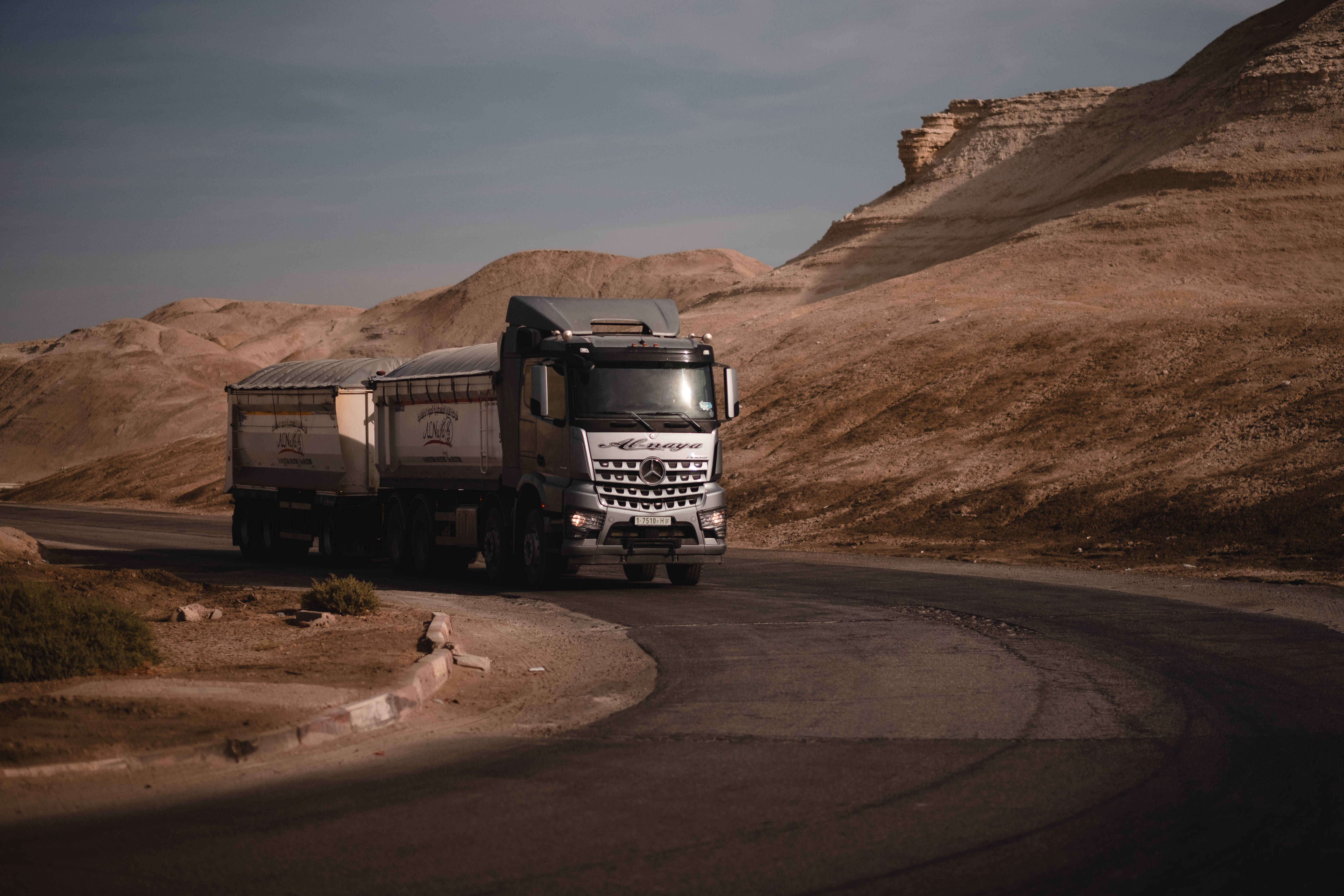 Truck 桌布 壁紙 — 一輛銀色 Mercedes-Benz 卡車在乾旱山丘環繞的蜿蜒沙漠道路上行駛，天空暗淡。