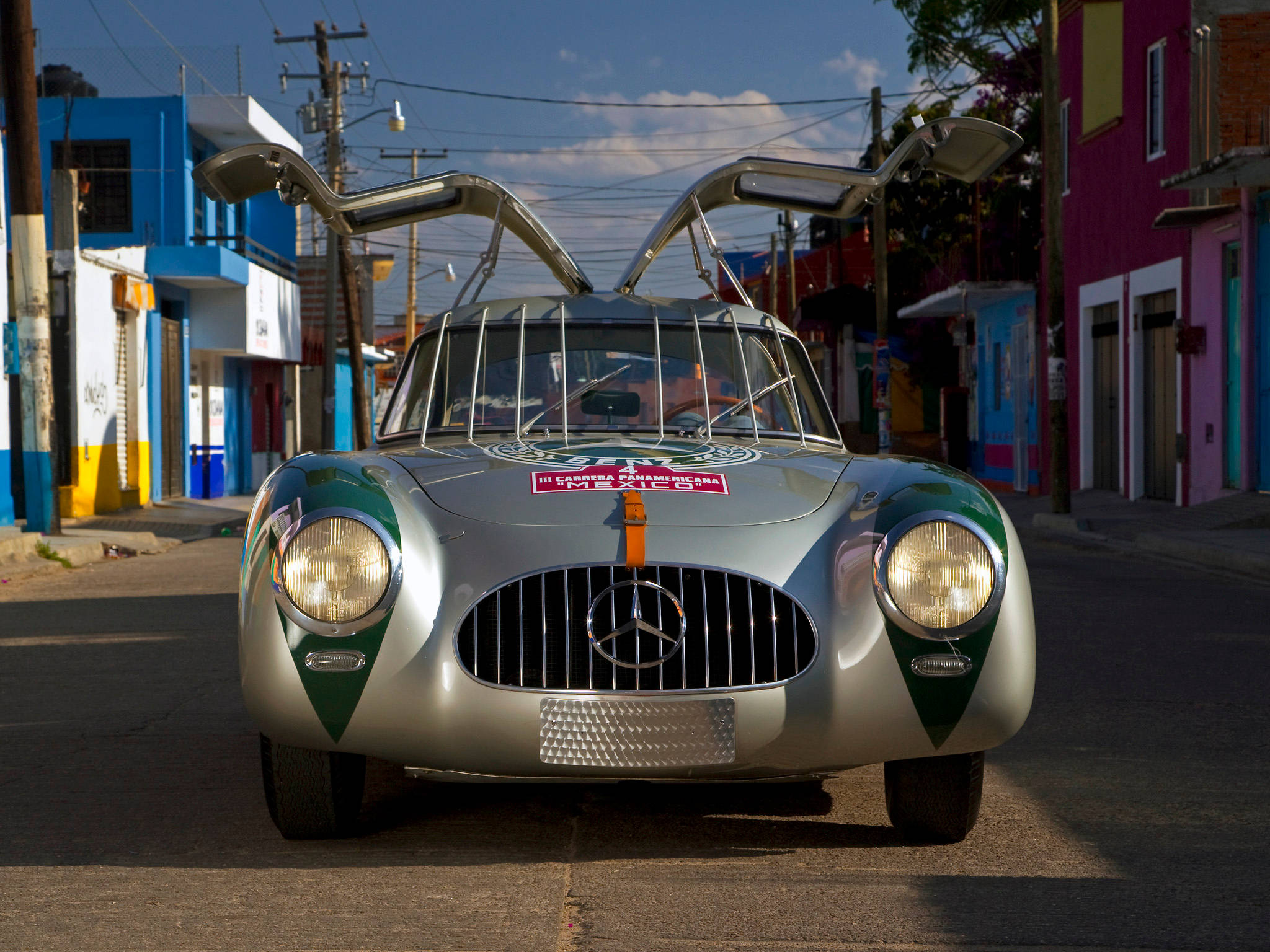 高畫質 賓士 桌布 壁紙 — 一輛銀色 Mercedes-Benz 300 SL 鷗翼門打開，停在色彩繽紛的街道上，兩旁是鮮豔的建築物，上方是明亮的藍天。
