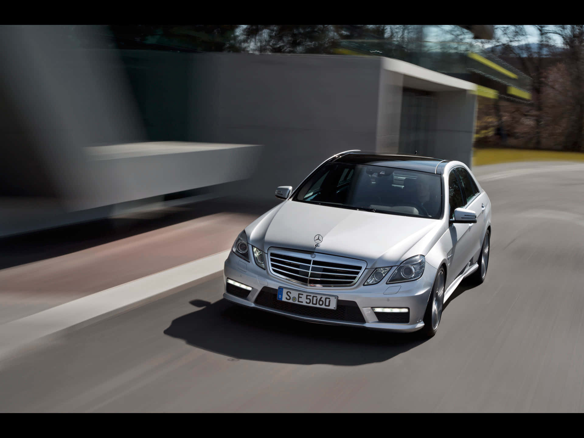 Mercedes Benz Clase E 桌布 壁紙 — 一輛銀色 Mercedes E-Class 轎車在現代化道路上疾馳，背景有模糊的動態和時尚建築。