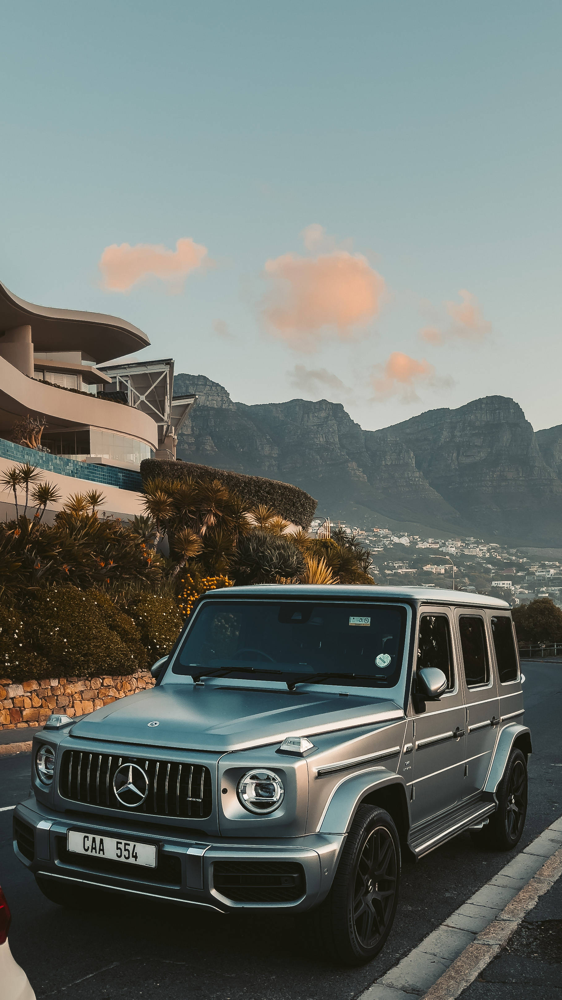 amg iPhone 桌布 壁紙 — 一輛銀色 Mercedes G-Wagen 停泊在沿海道路上，背景是現代建築和桌山，天空呈現柔和的日落色彩。