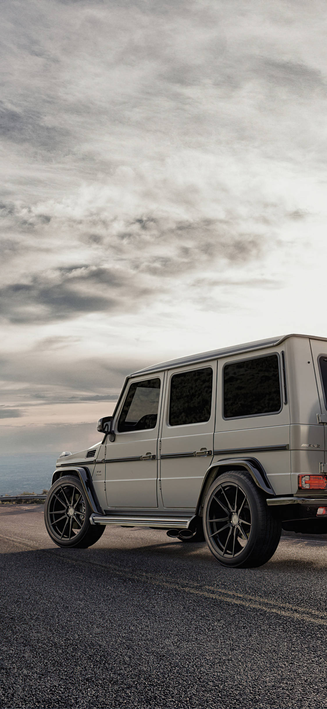 賓士 iPhone 桌布 壁紙 — 一輛銀色 Mercedes G-Wagen 停泊在海岸公路上，天空烏雲密布，氣氛戲劇化。