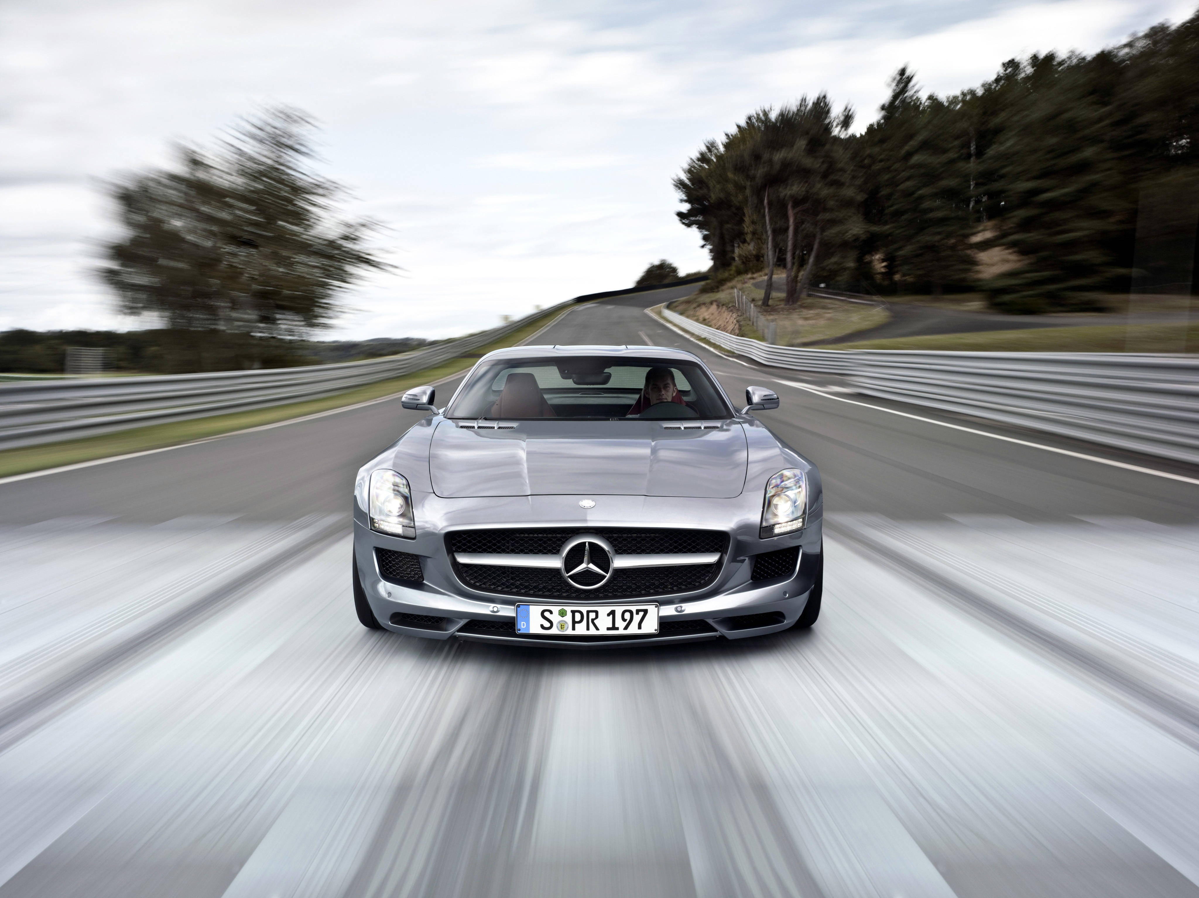 benz 4K 桌布 壁紙 — 一輛銀色 Mercedes SLS AMG 跑車在蜿蜒的道路上疾馳，背景有模糊的樹木和護欄。
