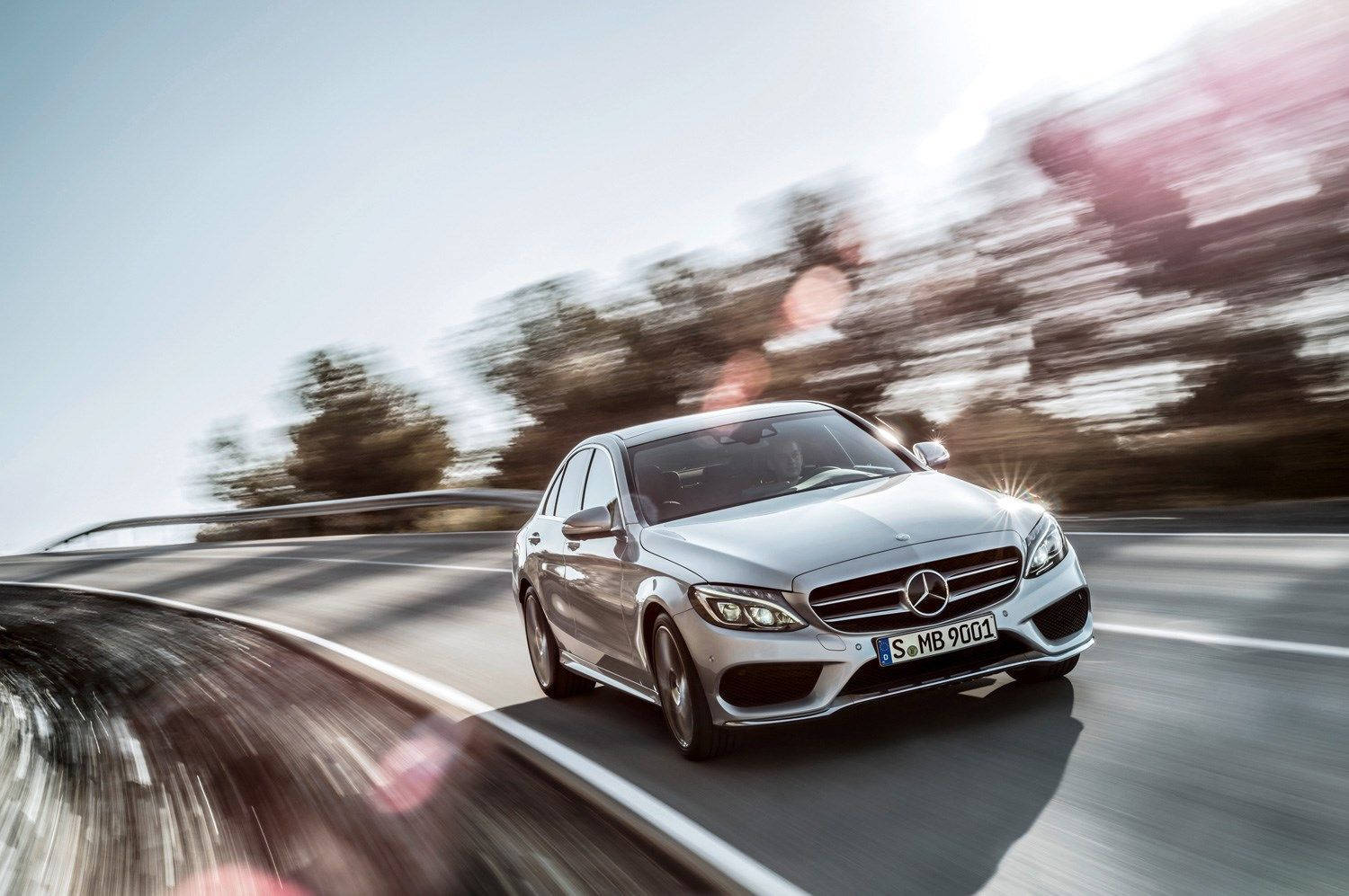 Mercedes Benz C300 桌布 壁紙 — 一輛銀色 Mercedes 房車在晴朗天空下，沿著蜿蜒道路疾馳，背景有模糊的樹木和明亮的鏡頭光暈。
