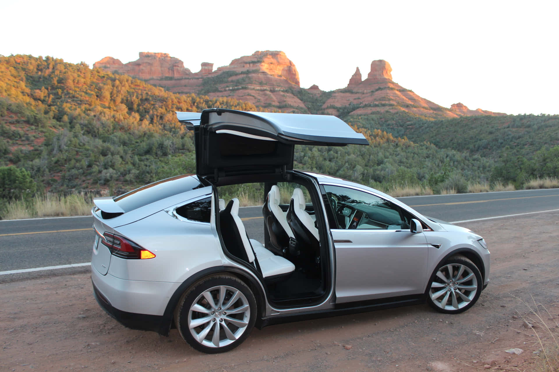 特斯拉 桌布 壁紙 — 一輛銀色 Tesla Model X 打開鷹翼門，停在沙漠公路上，背景是日落時分的紅色岩層。