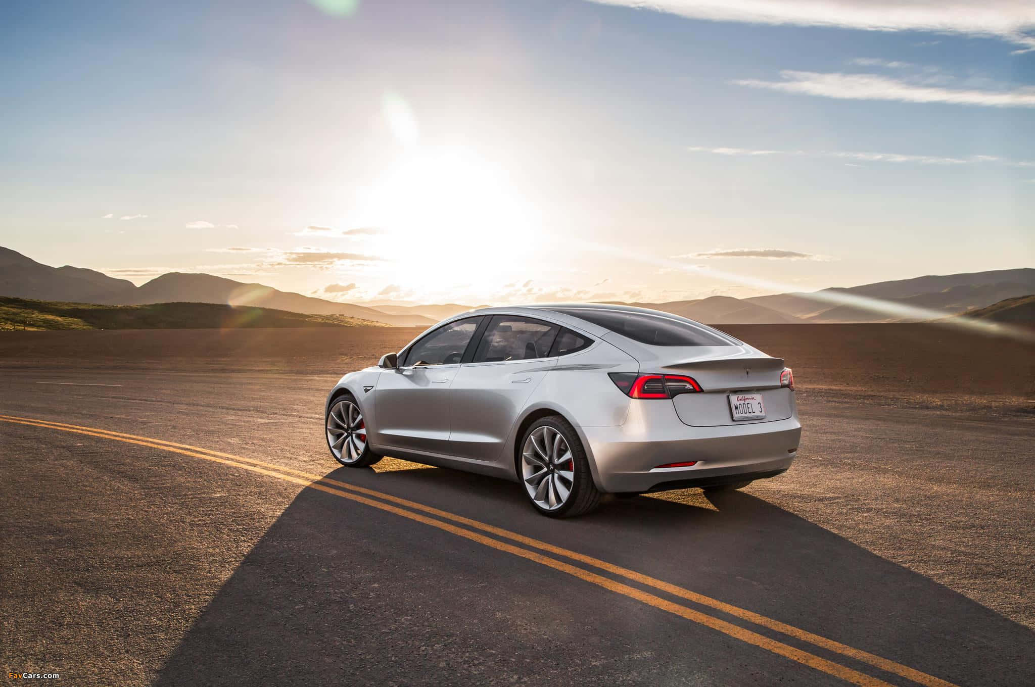 Tesla Model 3 桌布 壁紙 — 一輛銀色 Tesla Model 3 在日落時分沿沙漠道路行駛，背景有山脈。