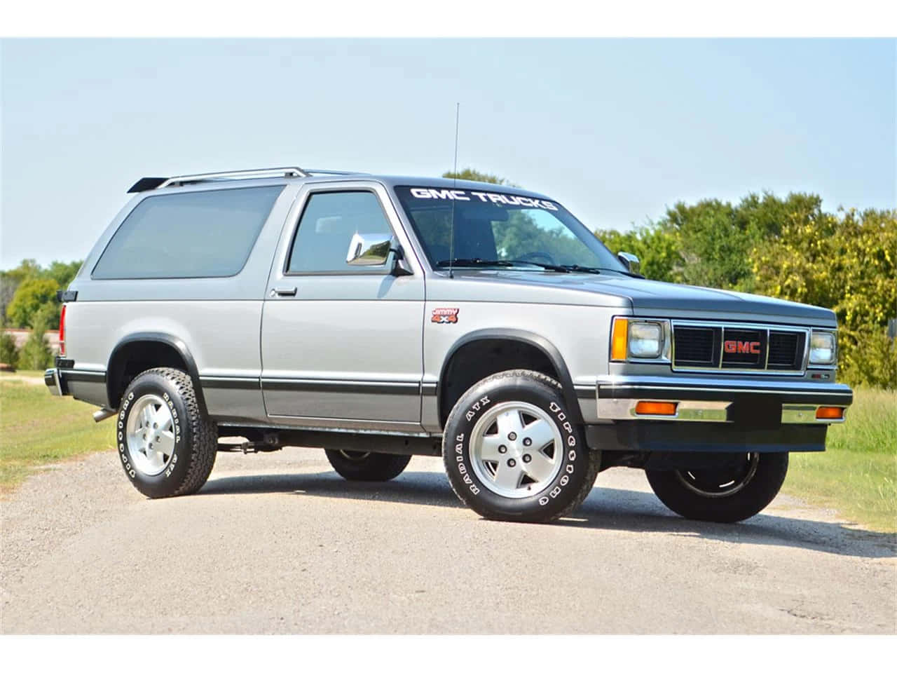 Gmc Jimmy 桌布 壁紙 — 一輛銀色1980年代GMC Jimmy SUV停泊在碎石上，背景有綠樹，天空清澈湛藍。