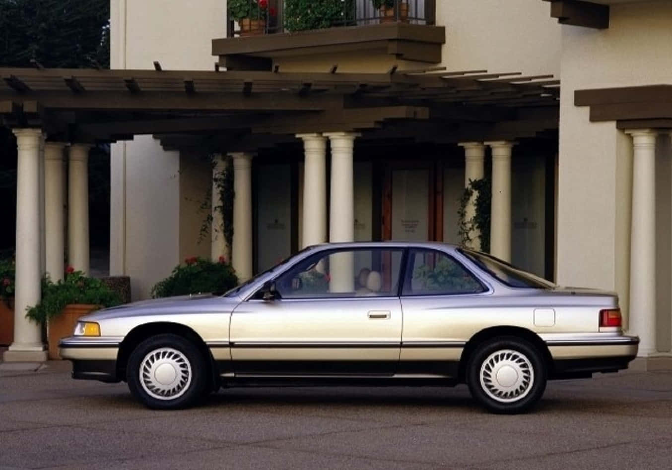 Acura Legend 桌布 壁紙 — 一輛銀色1990年代轎車停放在柱式涼棚下，前方是米色灰泥建築物，配有盆栽植物。