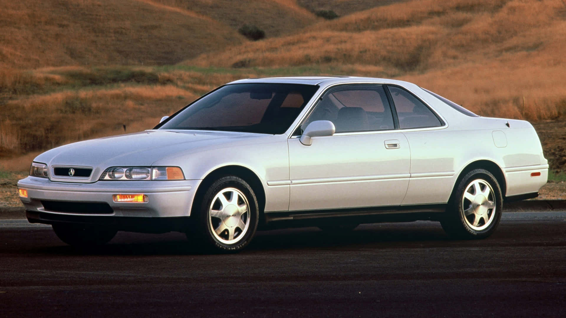 Acura Legend 桌布 壁紙 — 一輛銀色Acura Integra雙門跑車在日落時停泊在一條有金色山丘背景的道路上。