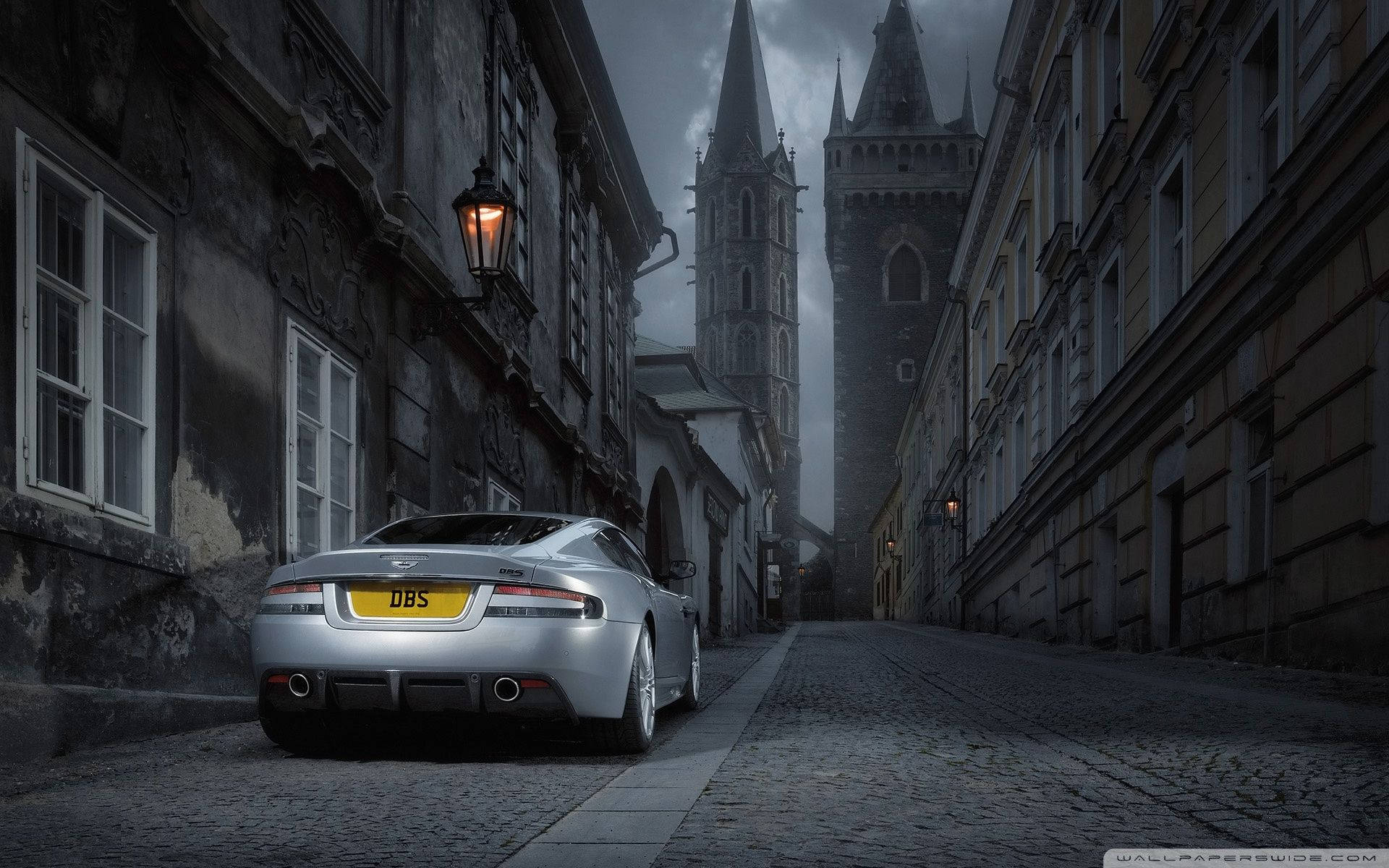 奧斯頓馬丁 桌布 壁紙 — 一輛銀色Aston Martin DBS停泊在鵝卵石街道上，兩旁是古老的歐洲建築，陰沉天空下遠處有教堂尖塔。