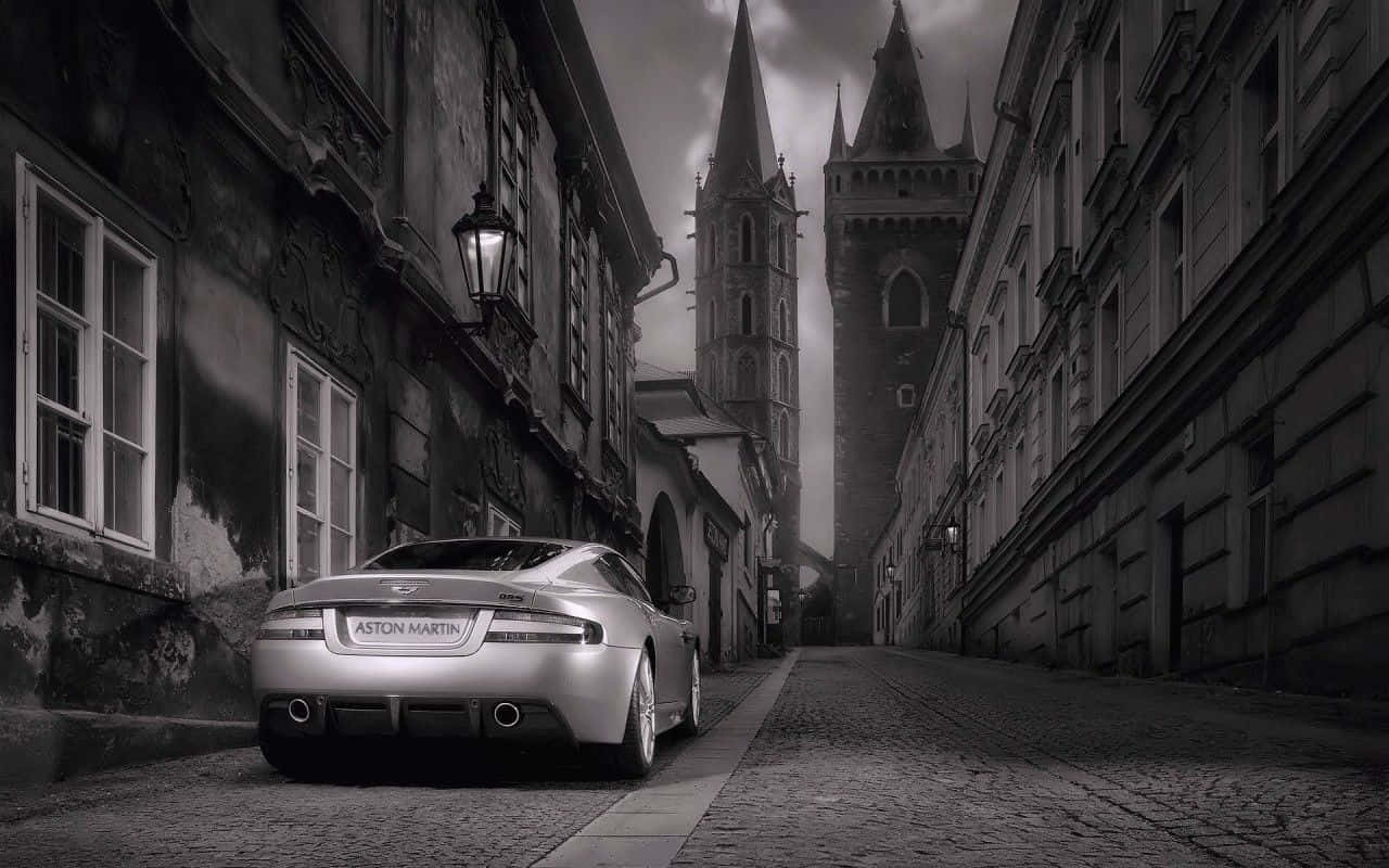 Black And White Car 桌布 壁紙 — 一輛銀色Aston Martin跑車停泊在鵝卵石街道上，兩旁是帶有哥德式尖頂的古老歐洲建築，天空陰沉多雲。